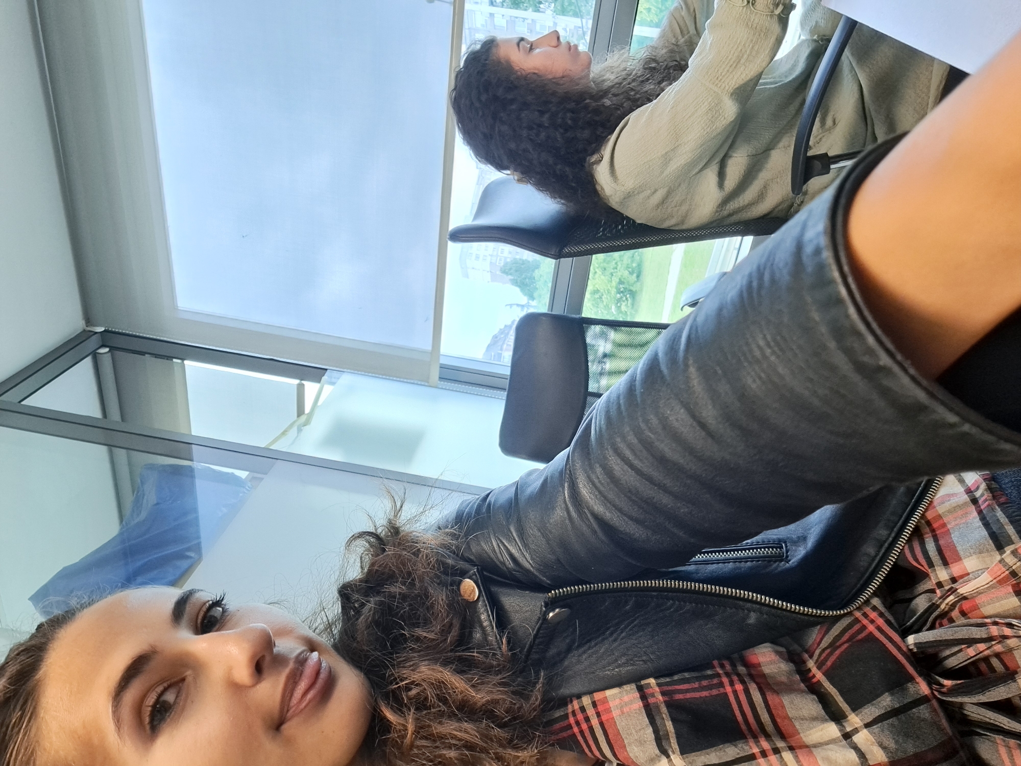 A selfie taken from a close-up perspective shows the face of a young woman with long, curly brown hair and brown eyes. She is wearing a black leather jacket over a red, black, and white plaid shirt. Her tongue is slightly sticking out of her mouth. In the background, another young woman with long, curly brown hair is seated at a desk in an office environment, facing away from the camera. She is wearing a light green textured top. Large windows with blinds are visible behind the seated woman, revealing a cityscape with buildings and trees. The environment appears to be a modern office with glass partitions and office chairs.