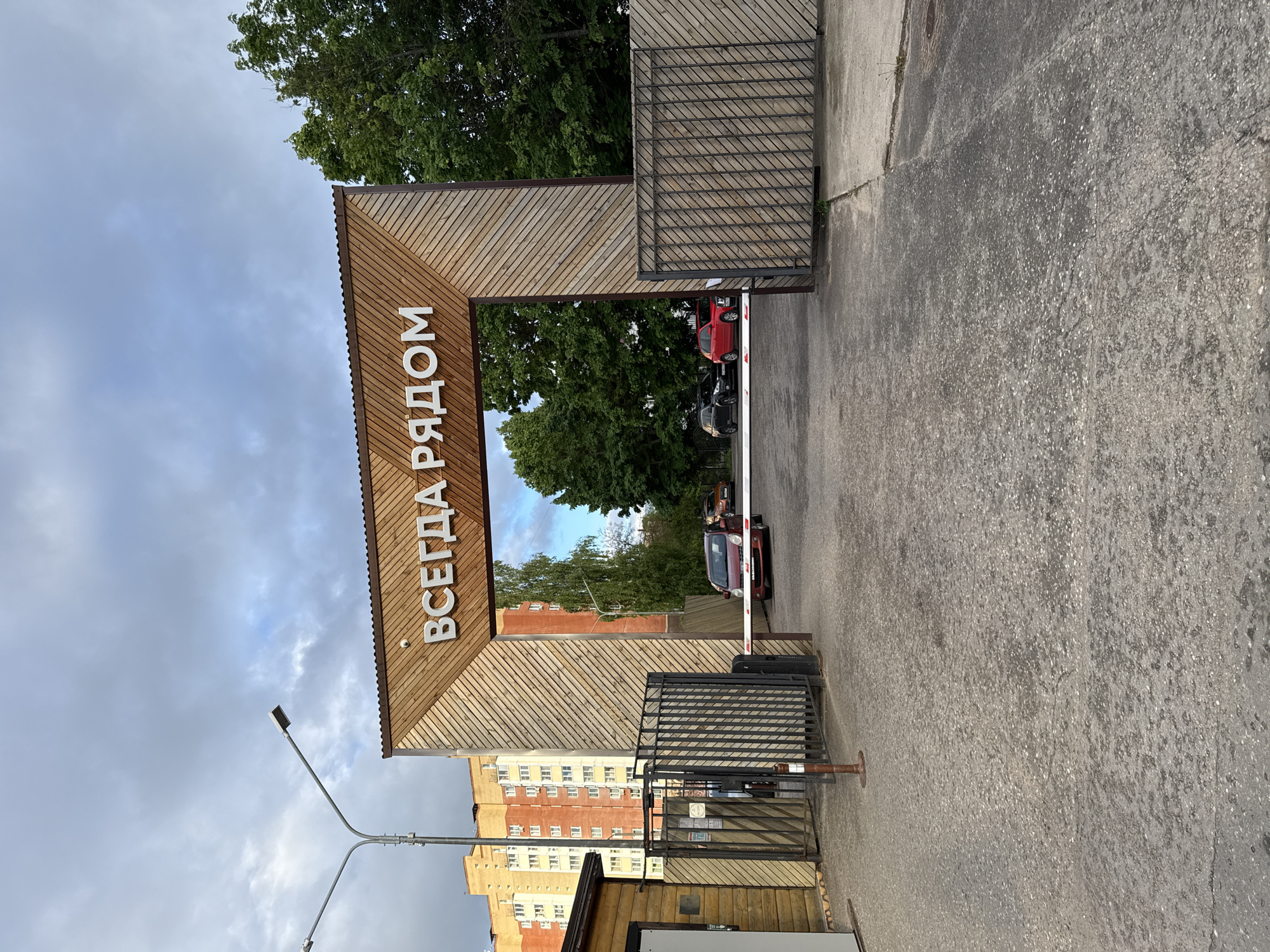 The image depicts the entrance to what appears to be a residential complex or gated community in Peresvet, Russia. A large wooden archway spans the entrance, bearing the inscription "ВСЕГДА РЯДОМ" in white letters. A metal gate and a security barrier are visible. A few cars can be seen beyond the barrier. A multi-story apartment building rises in the background to the left of the archway. A streetlight stands near the gate. The ground is paved with asphalt. The sky is overcast with clouds. Green trees are visible behind the barrier.