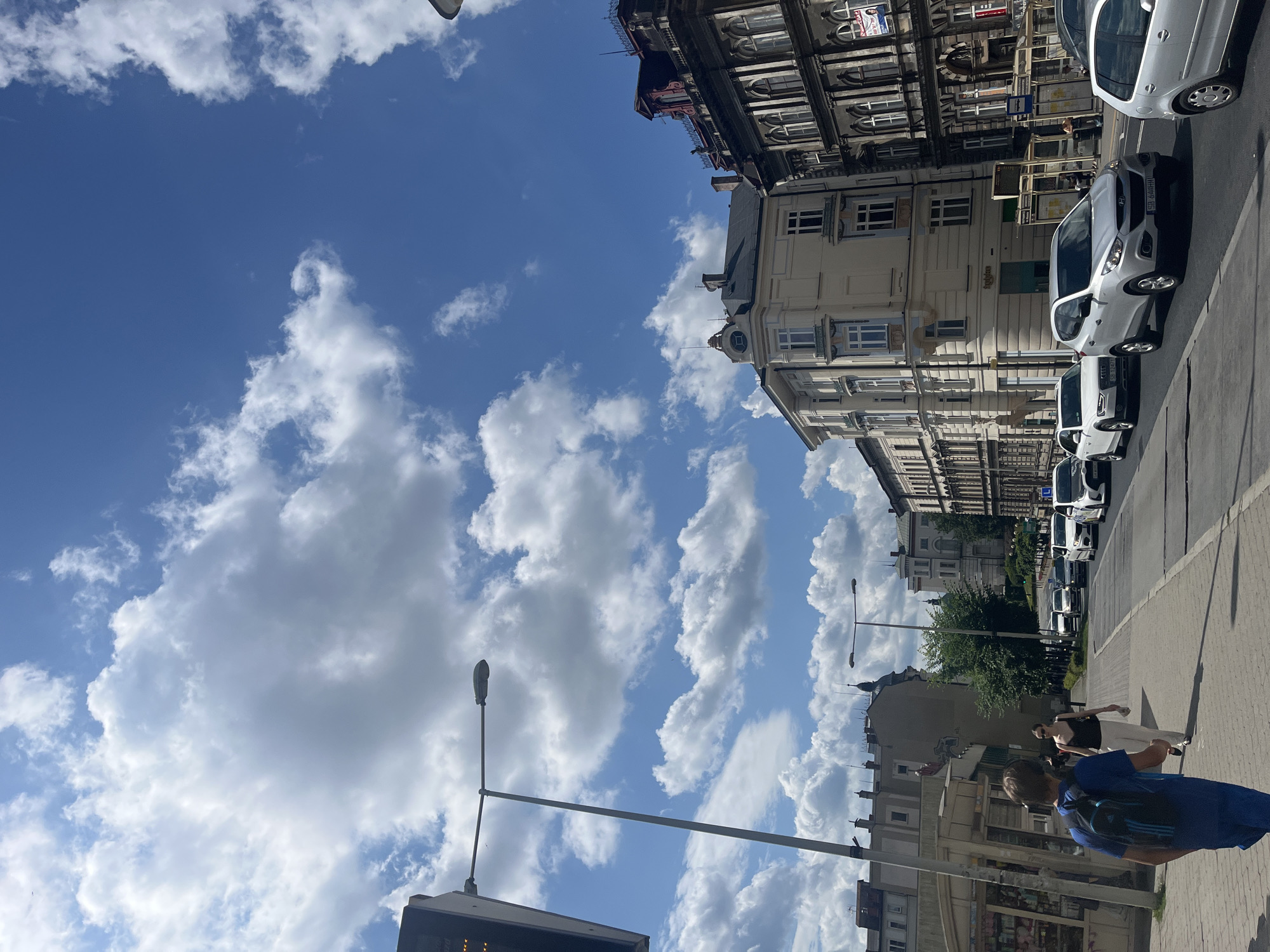 A street scene in an urban environment under a bright, cloudy blue sky. Several cars, including a silver Hyundai SUV and a white Audi sedan, are driving on the right side of the road. People are walking on the sidewalk on the left side of the road. The buildings lining the street feature classic architectural styles with multiple stories and windows. A lamppost stands on the sidewalk, and a kiosk is visible on the far left. The image captures a typical day in a city.