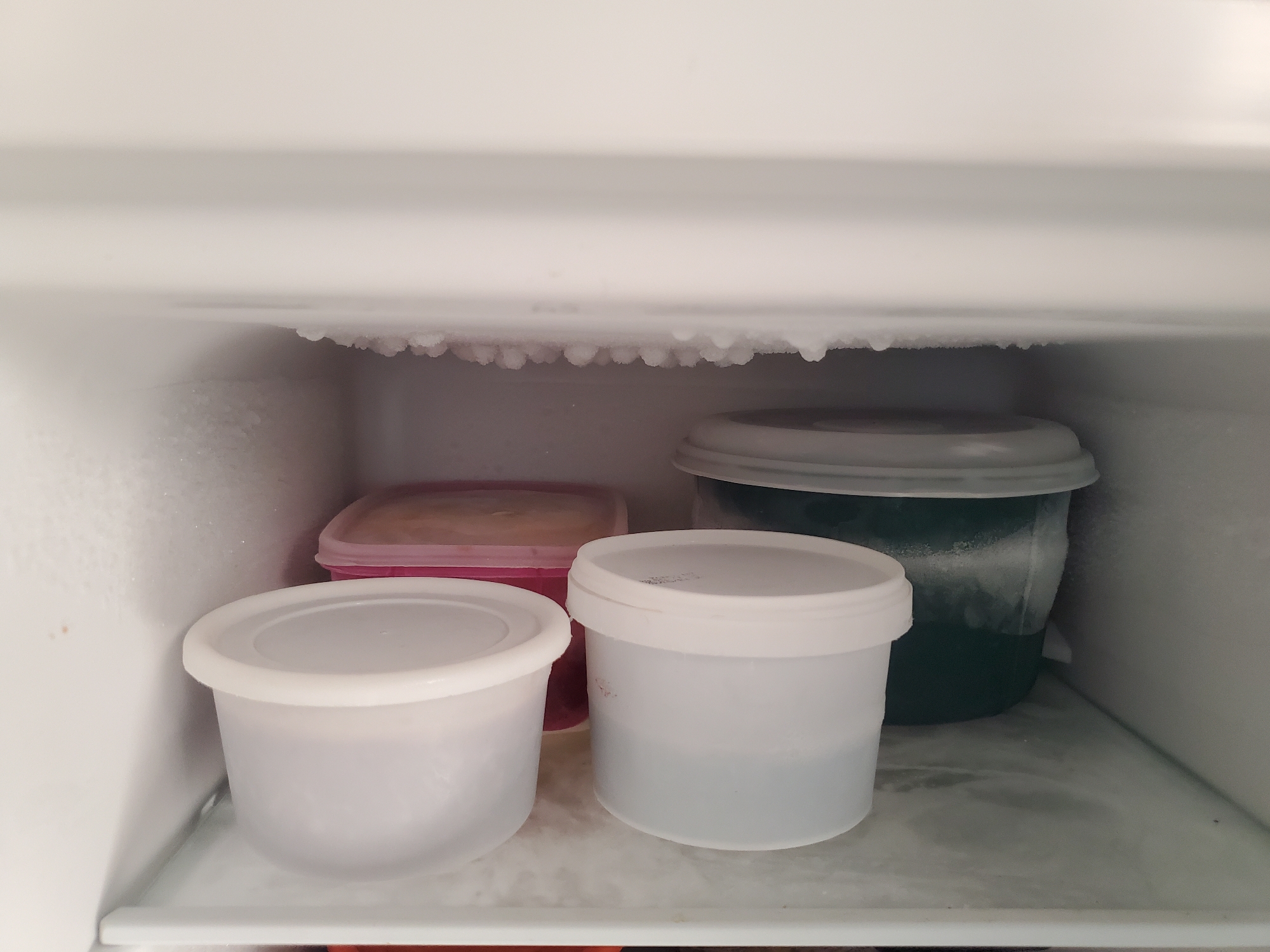 The image shows the interior of a freezer, likely in Barden Yana, Nigeria. Four containers of varying sizes and colors are visible on a shelf. The freezer walls and ceiling are covered with frost. The containers appear to hold frozen food or liquids. No people are visible. The environment is cold and indicates a functioning freezer.
