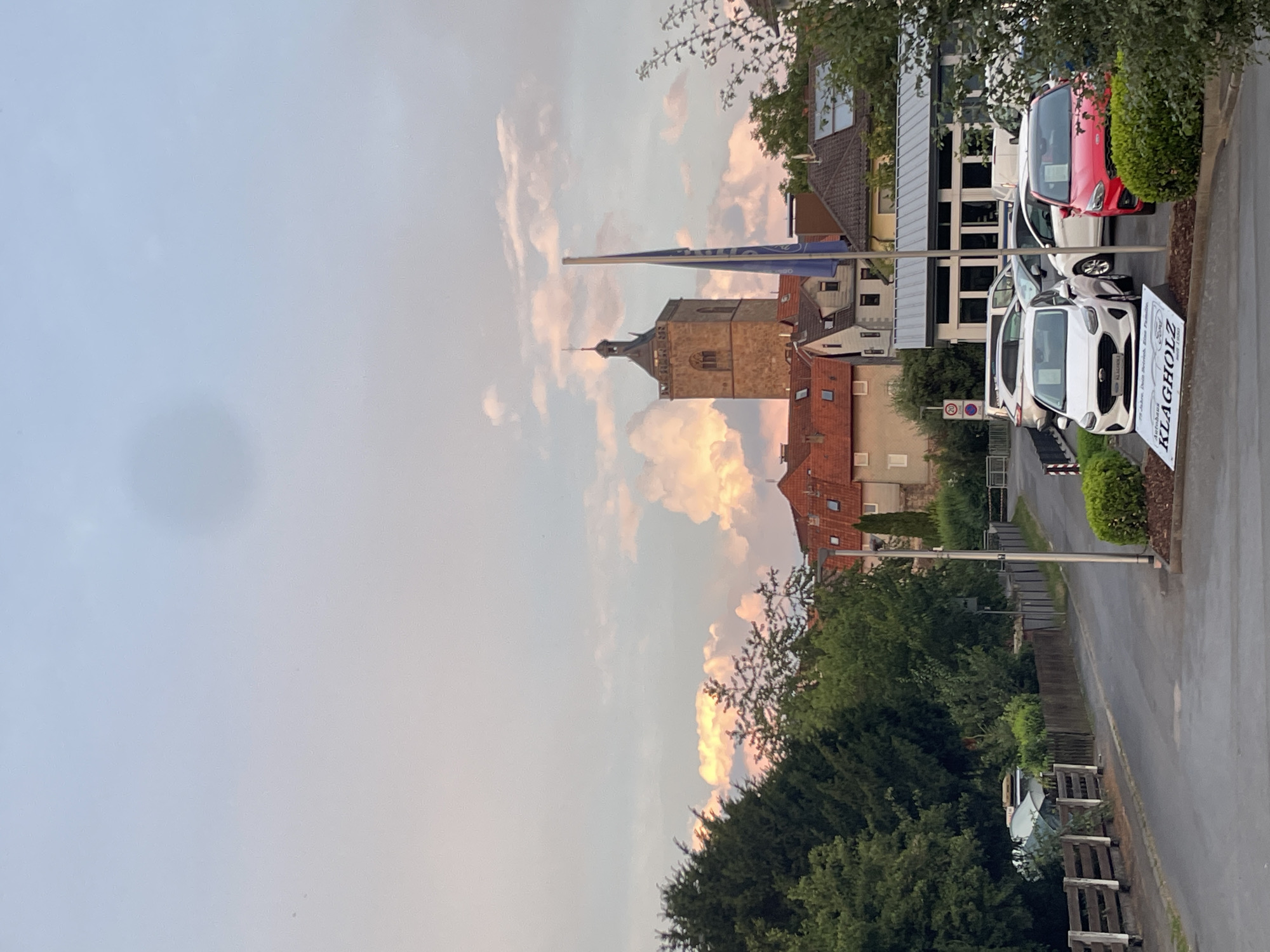 The image captures a view of a street in Neukirchen, Germany, with a church tower prominently visible in the background. The sky above is filled with soft clouds tinged with pink and orange hues, indicating it is either sunrise or sunset. In the foreground, several cars are parked on what appears to be a car dealership lot, with a white Ford and a red Ford being the most noticeable. A blue flag on a pole stands near the dealership. Trees line the left side of the street, and buildings with red-tiled roofs are visible behind the trees and around the church tower. A wooden fence runs along the left side of the road. The overall scene depicts a quiet street in a European town, with a blend of architectural elements and natural scenery.