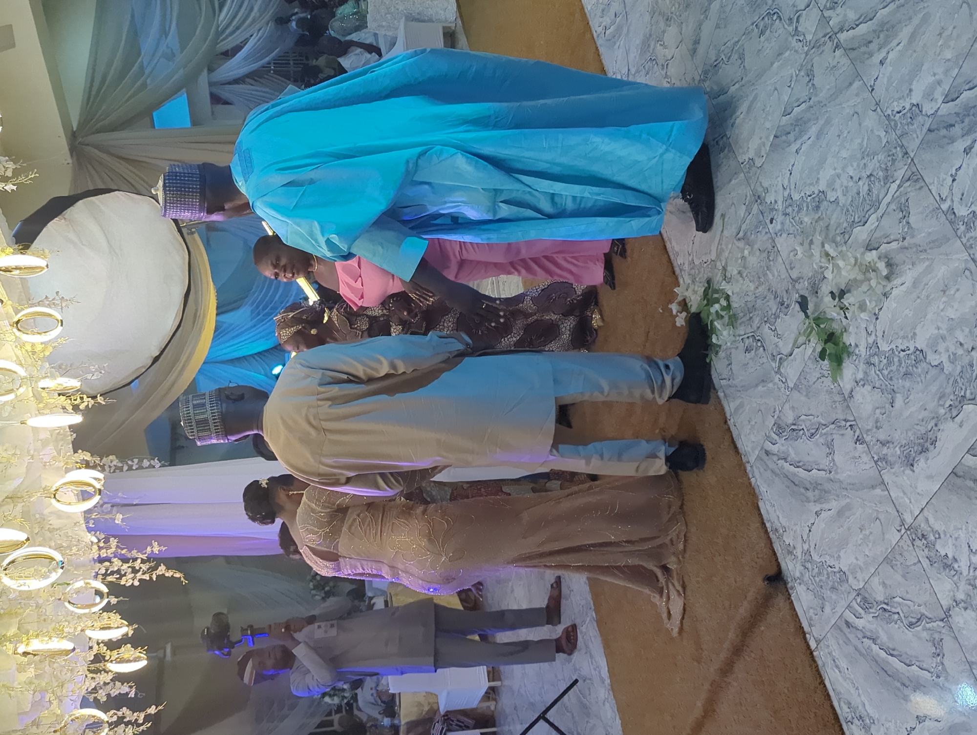 In Barden Yana, Nigeria, at an indoor event, four individuals are the primary focus. A person in a light gray traditional outfit is holding a camera with a gimbal, facing left, in the background left. To their right, a woman in a floor-length beige sequined dress is looking forward, with a person in a light gray traditional outfit standing behind her, partially obscuring her. In the foreground right, a person in a bright blue traditional outfit faces away from the camera. To their left, a woman in a pink off-the-shoulder dress and a woman in a patterned dress are visible. The environment features decorative hanging lights from the ceiling, draped fabric backdrops, and a combination of a beige carpet and marble-tiled flooring. Scattered white flowers are on the floor in the foreground right.