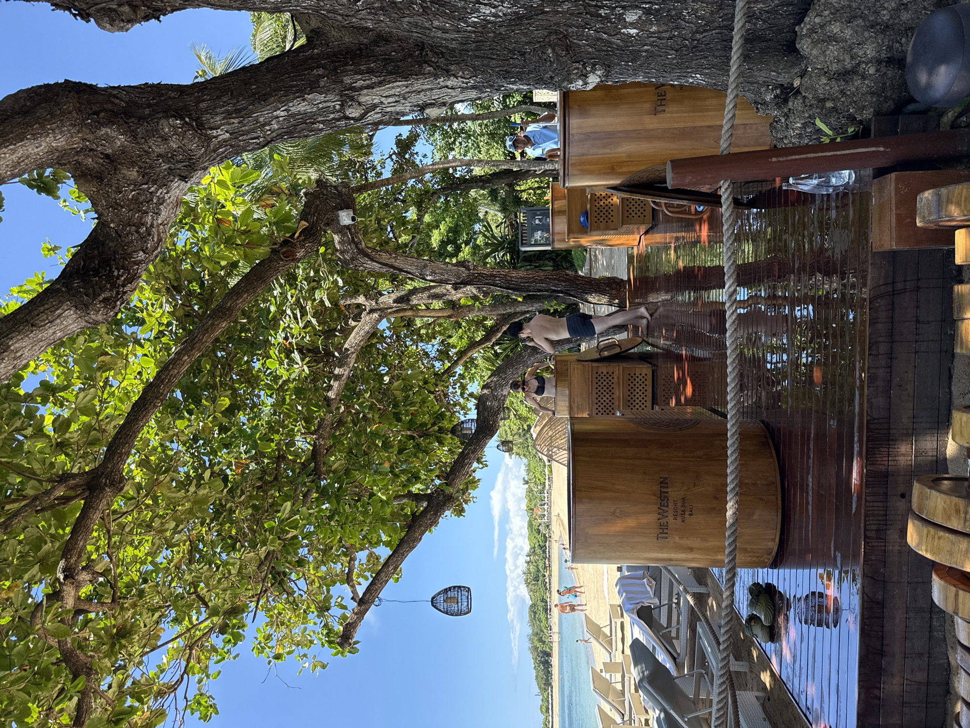 The image shows an outdoor scene in Nusa Dua, Indonesia. A large tree with green leaves dominates the left side of the frame, partially obscuring the sky. To the right, wooden structures are visible, including cylindrical containers labeled "THE WESTIN NUSA DUA BALI". One man is bending over near one of these containers, possibly washing. A second person can be seen in the background. A beach is visible in the distance, with lounge chairs and several people on the sand and in the water. Hanging decorations are present in the tree branches. A rope runs vertically through the scene. Shoes are located on a wooden deck.