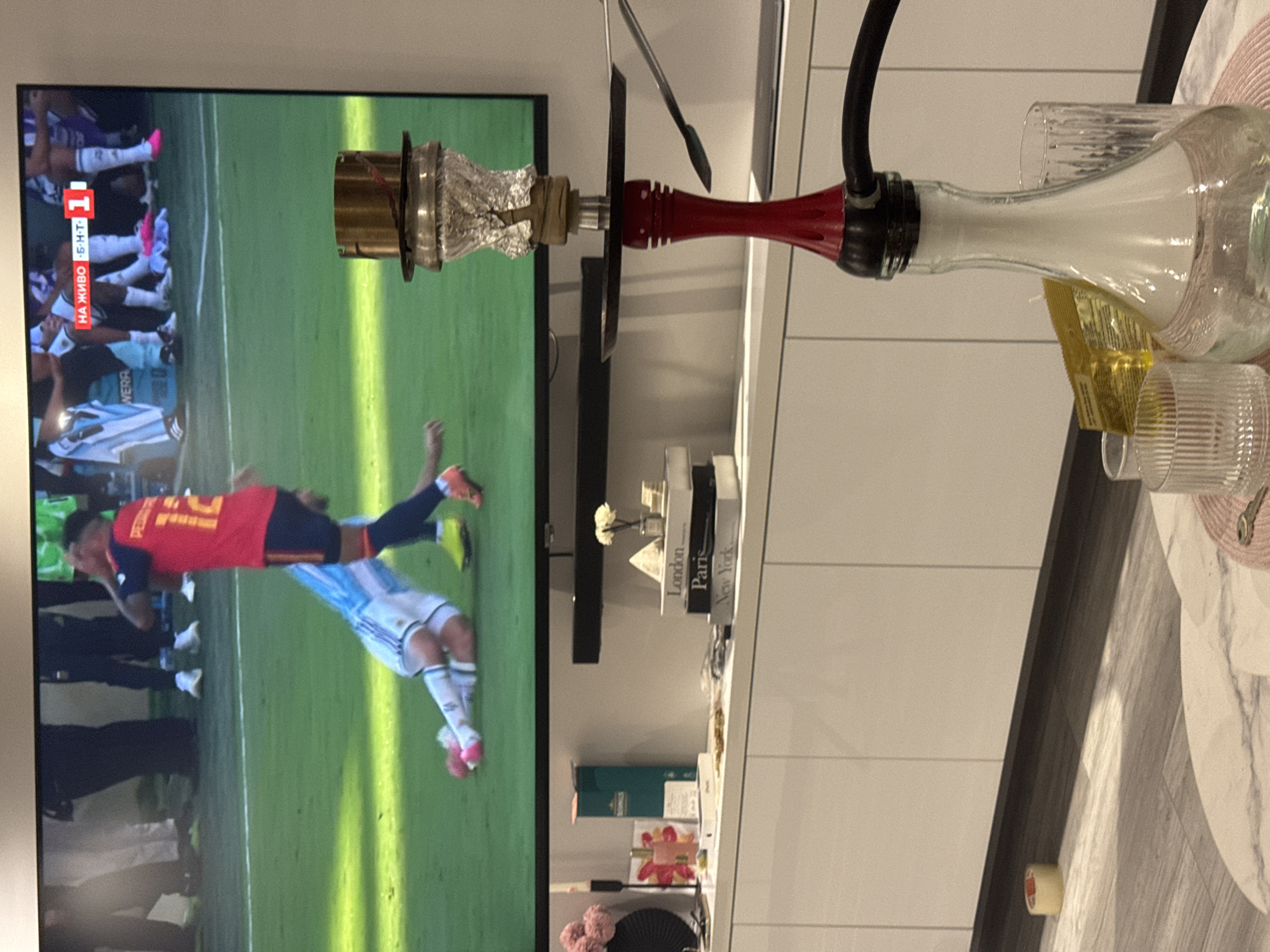 The image depicts an indoor scene with a large television screen dominating the upper portion, displaying a live football match. The screen shows two players on a green field, one in a red jersey with the number 12, and another in a blue and white striped jersey lying on the ground. A scoreboard overlay on the TV indicates "НА ЖИВО Б.Н.Т. 1". In the foreground, a red and black hookah is prominently featured, with its glass base filled with water and smoke. The hookah is placed on a white marble-patterned table alongside two clear glasses and a small package. Behind the table, a white credenza holds several decorative items, including books with titles like "London," "Paris," and "New York," a small vase with flowers, a candle, and a bottle of perfume. The room appears to be a living space, with a floor and wall visible. There is no specific indication of the city of Burgas, Bulgaria, in the image itself.