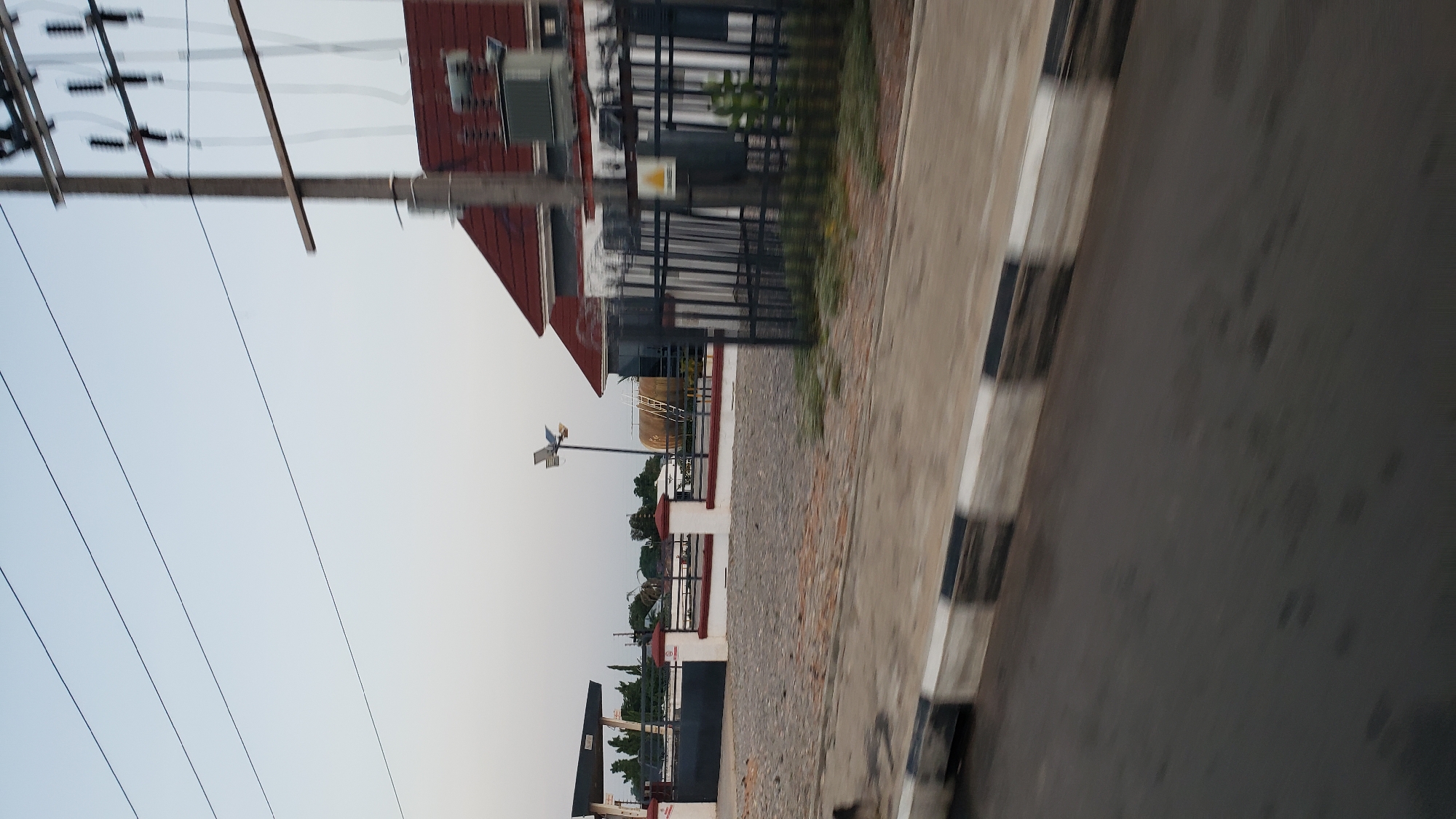 This is a medium-shot, eye-level, outdoor photograph of a street scene in Jalingo, Nigeria, taken in the early evening. The image captures a roadside view with a building and a power transformer. On the left, a building with a canopy structure, likely a gas station or service station, is partially visible. A black metal gate encloses an area filled with gravel. Red and white bollards with a dark red railing line the perimeter. In the background, beyond the gate, are trees and more buildings. A tall pole with a solar-powered lamp stands near the center of the frame. To the right, a modern building with a red roof is visible. A tall electrical pole stands in front of this building, supporting power lines and insulators. A large, grey electrical transformer is mounted on a concrete base next to the pole. A black metal fence with vertical bars, topped with barbed wire, encloses the property. A yellow triangular warning sign with a black lightning bolt symbol is attached to the fence. The foreground shows a paved road with a curb marked with alternating black and white rectangular patterns, indicating a pedestrian crossing or hazard marking. The road surface appears wet and reflects the sky. The sky is a clear, pale blue, suggesting the end of the day or early morning. The image has a slight motion blur, indicating it was taken from a moving vehicle.