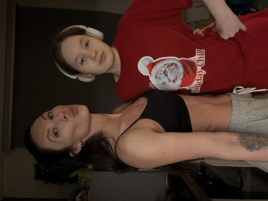 Two people pose for a photo. The person in the foreground is an adult woman in a black spaghetti strap tank top and gray sweatpants. She is lying down and looking at the camera with her lips puckered. Behind her is a younger person wearing a red t-shirt with a cat wearing a Santa hat on it. They are wearing headphones and looking at the camera.