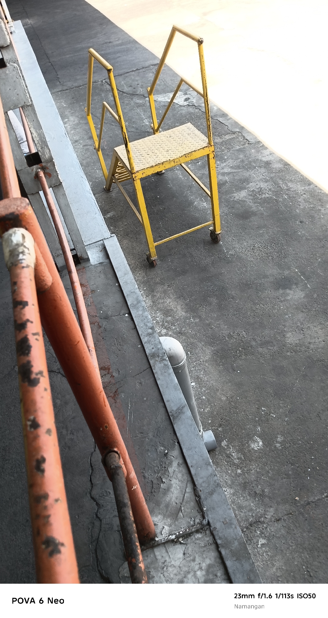 A yellow stepladder with a diamond-plate platform and wheels is positioned on a concrete surface. To the left of the stepladder, a rough, orange-painted metal railing is visible. The environment appears to be an outdoor industrial or service area, with a shadow cast across the concrete from an unseen object. The lower right corner of the image displays text indicating camera settings: "23mm f/1.6 1/113s ISO50" and the location "Namangan".