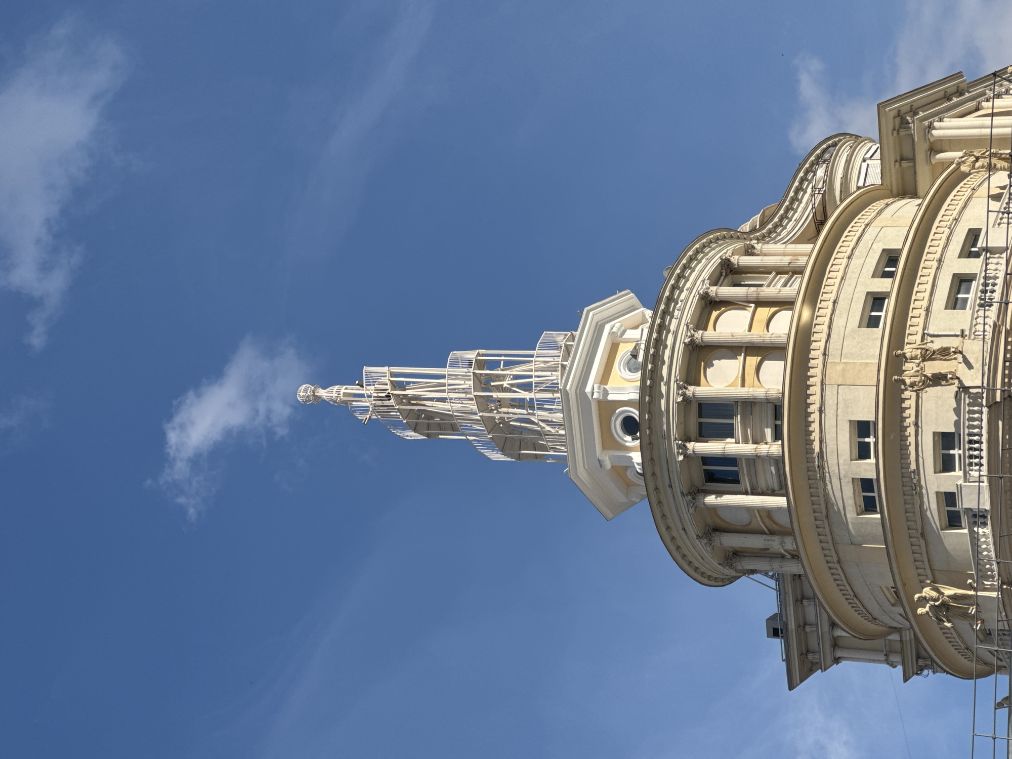 The image shows the top portion of a cream-colored, circular building against a bright blue sky with wispy clouds. The building features classical architectural elements, including columns, arched windows, and ornate detailing. A spiral-like white spire crowns the building. Statues of figures in classical attire are visible on balconies. The building's facade is detailed with multiple windows and decorative moldings. The location is the Moscow State University.