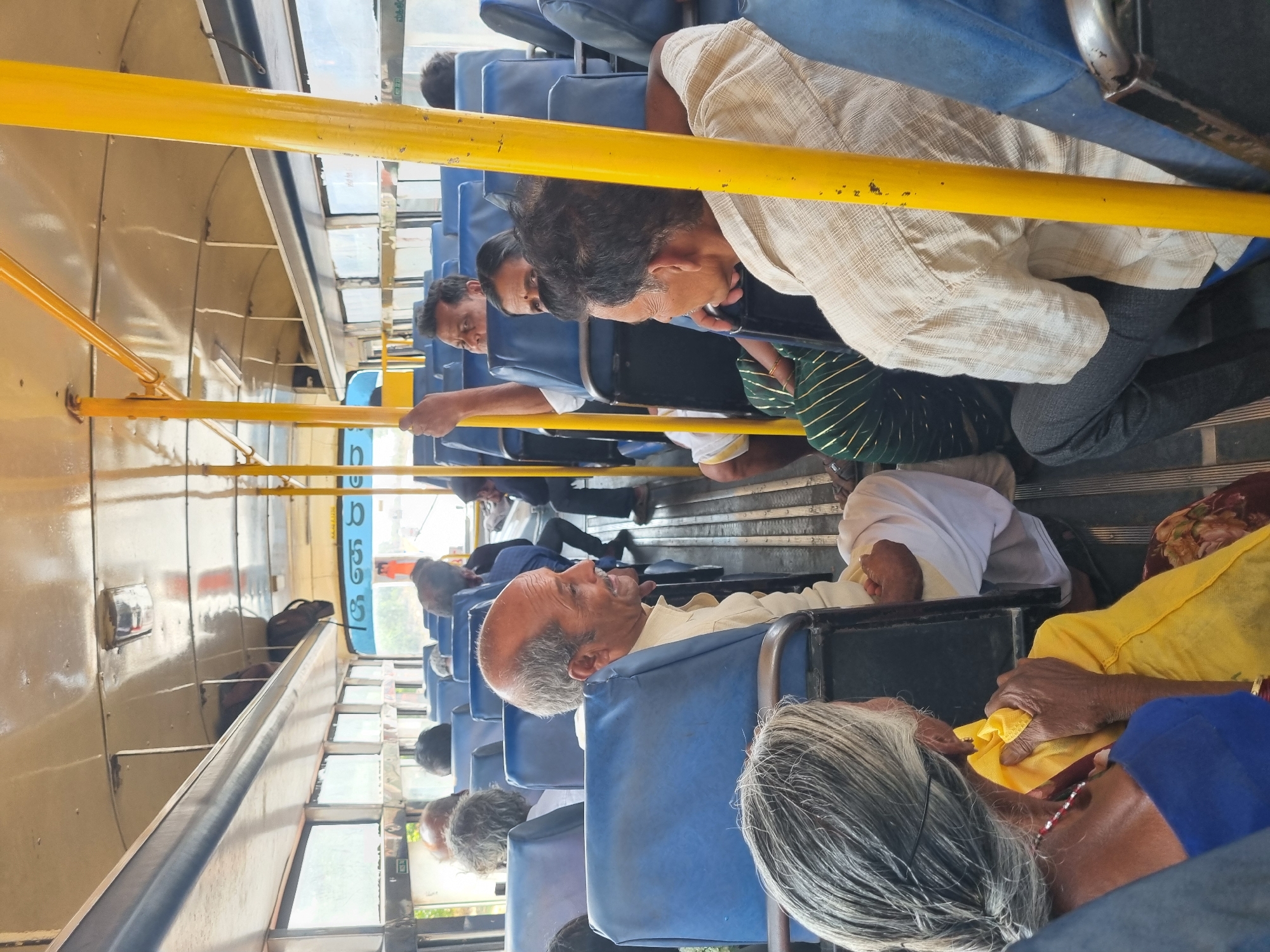 Inside a bus in Kodikonda Check Post, India, several passengers sit on blue seats, holding onto yellow handrails. The passengers include elderly individuals with gray hair, one wearing a white head covering, and others in everyday attire. Some are looking down or sideways, with the bus interior revealing metal panels and window views.