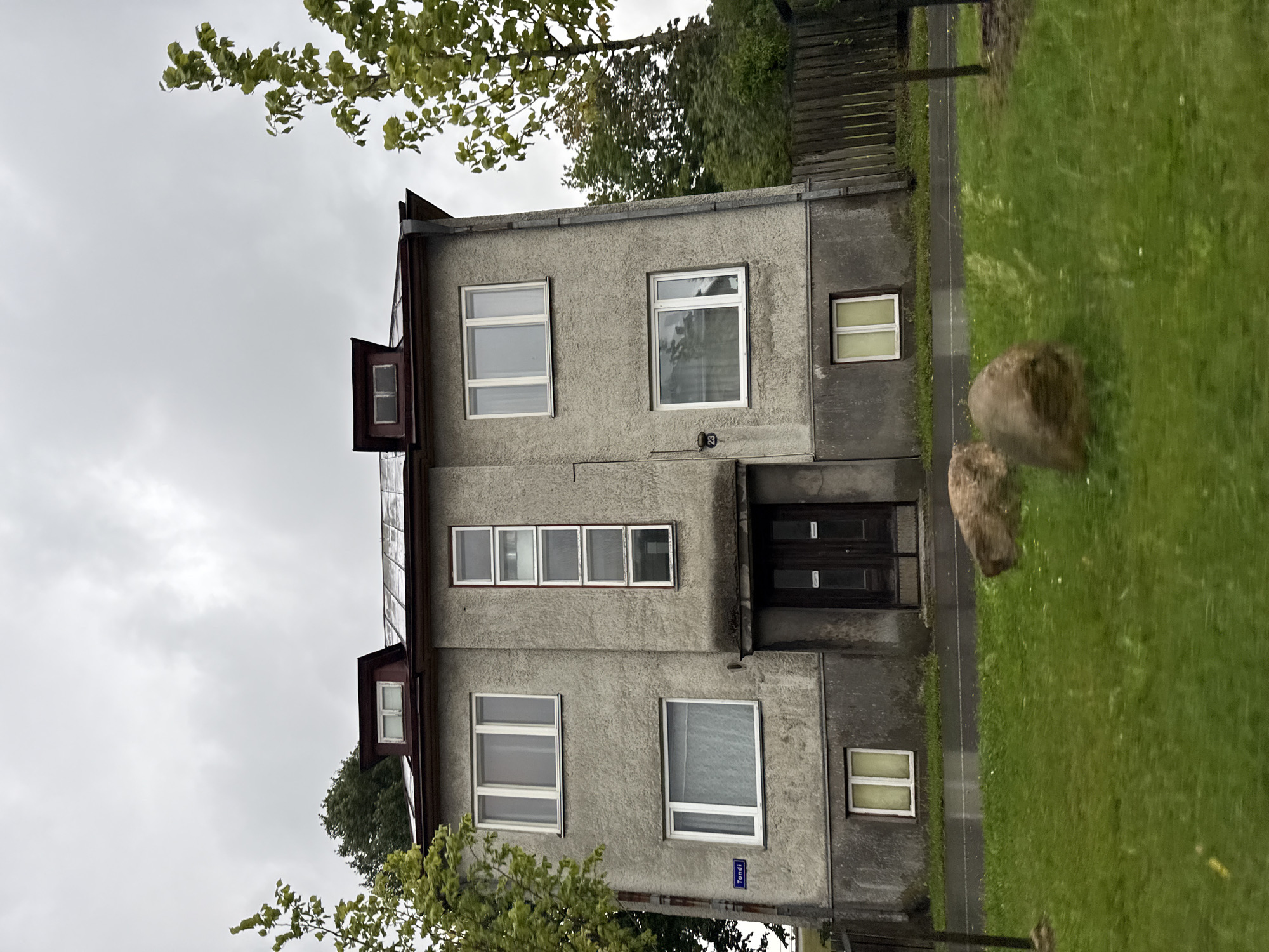 The image shows a three-story apartment building with a gray, stucco facade. The building has a dark-colored roof and two dormer windows on the top floor. There are several windows on each floor, some of which are covered with white curtains. The building appears to be located on a street in Tallinn, Estonia, as indicated by a street sign that reads "Tondi". In front of the building is a grassy lawn with two large rocks. The sky is overcast and the ground appears wet, suggesting it may have recently rained.