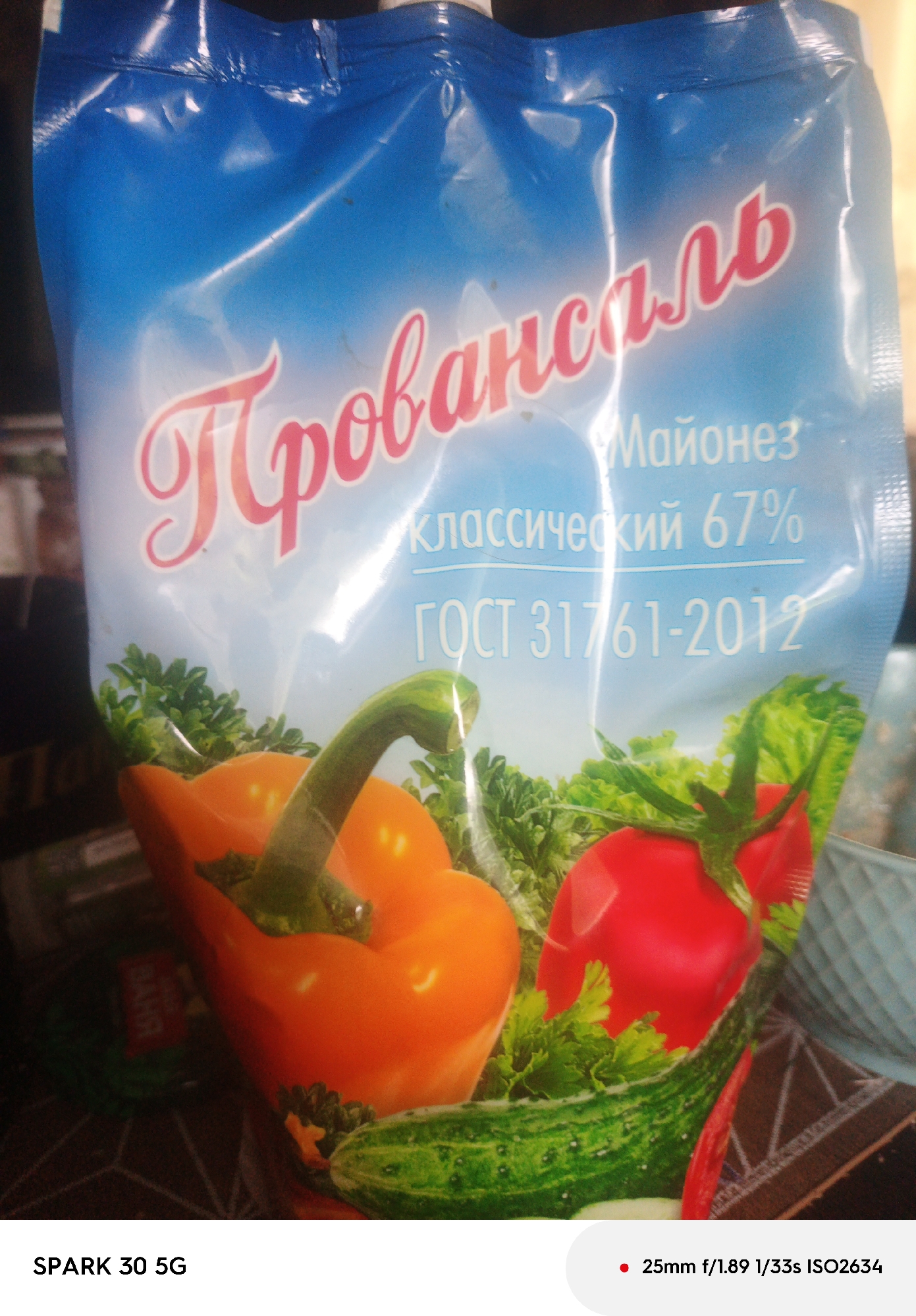 A front-facing, slightly angled medium shot captures a blue plastic pouch of mayonnaise with a printed image of vegetables. The top of the pouch is wrinkled and sealed. The product name, "Провансаль" (Provansal), is prominently displayed in pink, stylized cursive lettering across the upper portion of the pouch. Below this, in smaller white text, are the words "Майонез" (Mayonnaise), "классический 67%" (classic 67%), and "ГОСТ 31761-2012". The lower half of the pouch features a realistic printed illustration of vegetables, including a bright orange bell pepper, a red tomato, and green cucumbers and parsley. The background is dark and out of focus. In the bottom left corner of the image, the text "SPARK 30 5G" is visible. To the right of this text, a red dot is followed by camera settings: "25mm f/1.89 1/33s ISO2634". There are no people or animals present in the image, and the setting is indoors, likely a home environment, with no discernible connection to the city of Irsha, Russia.