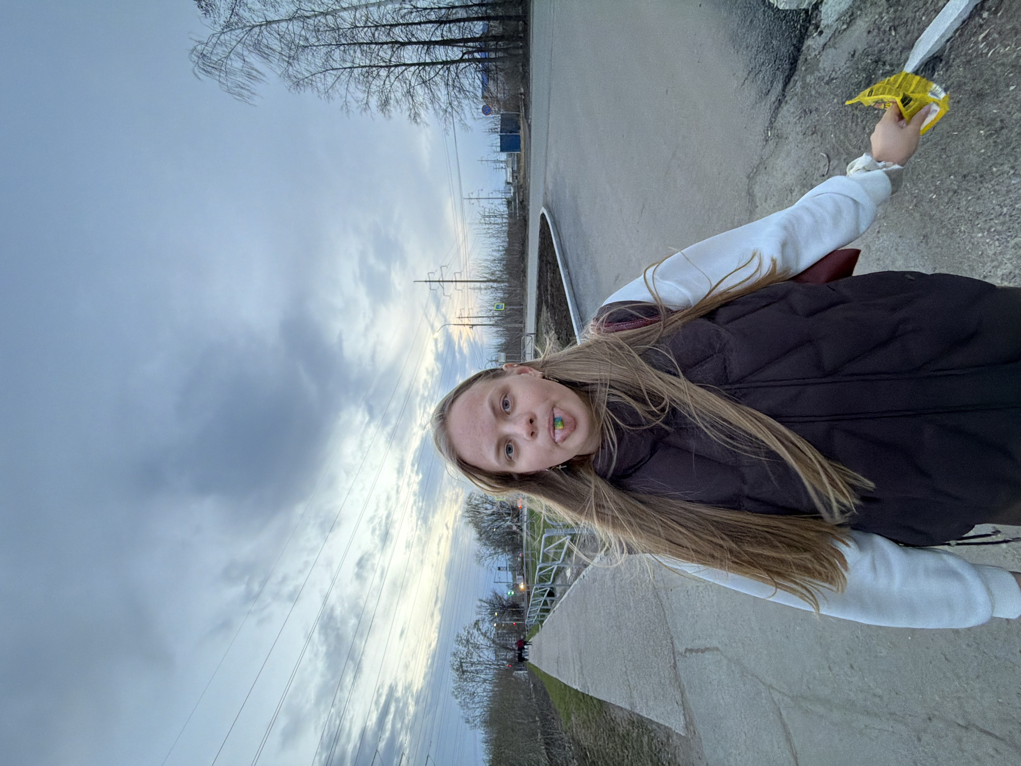 The image, originally oriented 90 degrees counter-clockwise, portrays a young woman outdoors in what appears to be Chaikovsky, Russia. She is shown from the waist up, with long blonde hair, looking directly at the camera. A small, colorful item, likely a piece of candy or gum, is visible in her mouth, giving her a slightly playful expression. She is wearing a white long-sleeved top beneath a dark, possibly quilted, vest. In her right hand, she holds a crinkled yellow package, possibly containing snacks or candy.
The setting is an urban or semi-urban environment with an overcast sky, showing patches of brighter light, suggesting diffused daylight, possibly late afternoon or early morning. Bare trees line the distant background, indicating a season like autumn, winter, or early spring. Power lines and utility poles are prominent features stretching across the midground.
To her left, a paved road or path is visible, bordered by a curb. Further into the background, a metal railing can be seen. Two distinct traffic signs are present: a blue circular sign with a white arrow pointing right, and a yellow diamond-shaped sign featuring a pedestrian symbol, indicating a pedestrian crossing. The ground consists of concrete or asphalt surfaces mixed with patches of sparse vegetation. No other legible text is visible on objects in the scene.