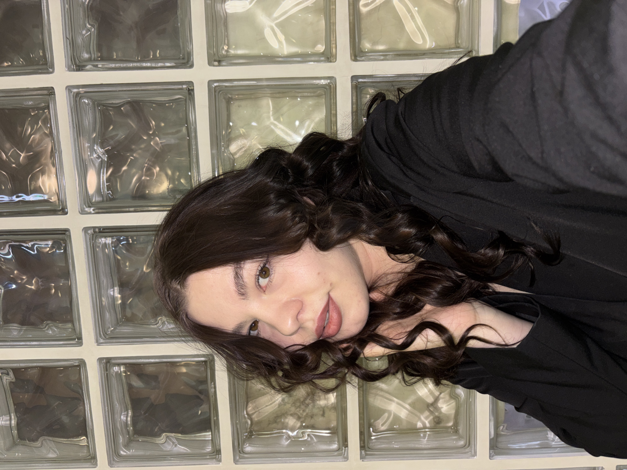 A woman with dark curly hair is seen in a close-up shot, likely a selfie, with her head resting on her hands. She is wearing a black top. Behind her is a wall of glass blocks. While I cannot pinpoint the exact location in Perm, Russia, the glass block wall suggests an interior space, possibly a building constructed in the late Soviet era.