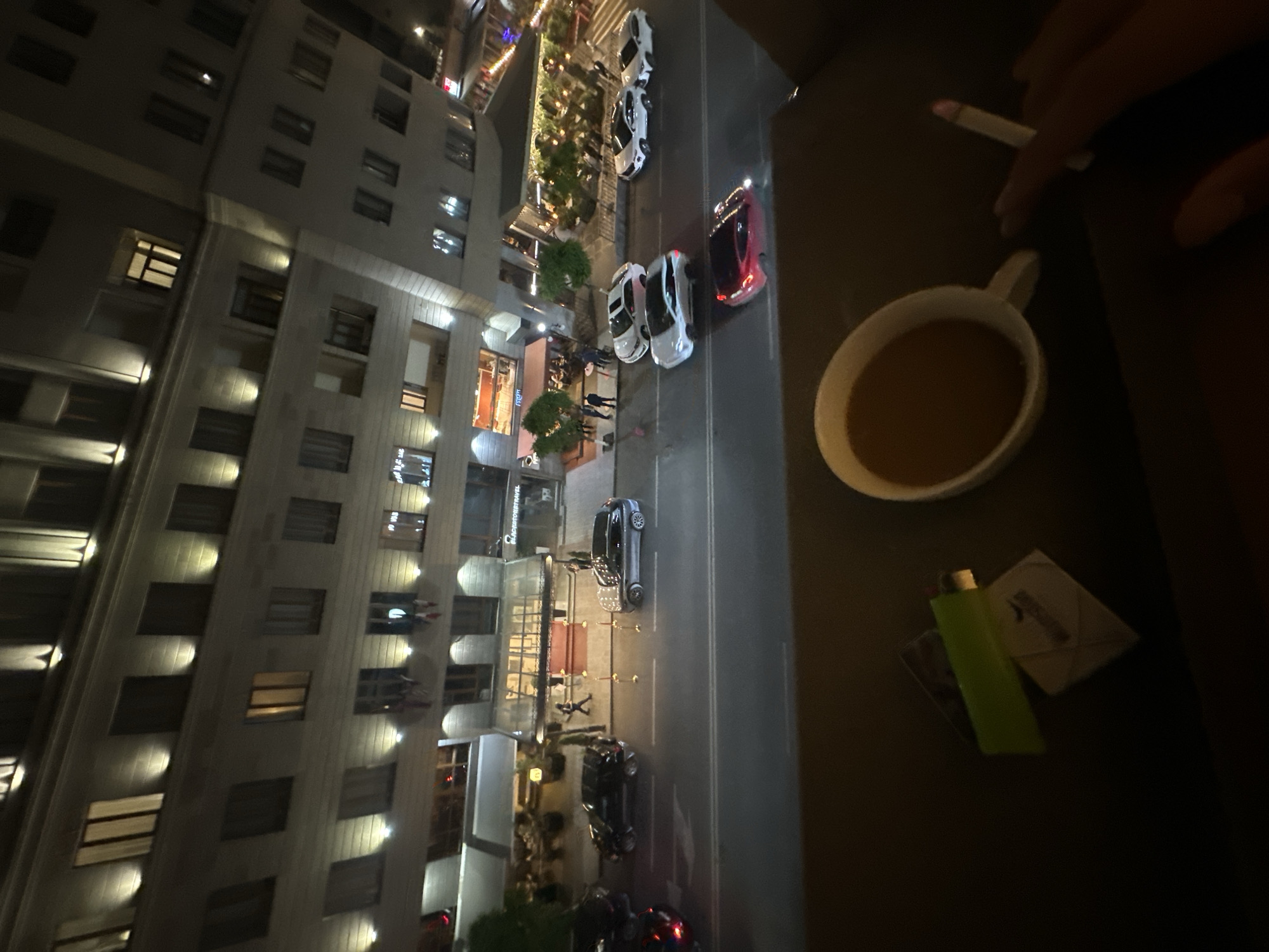 The image, taken from a high vantage point, shows a nighttime scene in Yerevan, Armenia. A tall, multi-story building with lit windows occupies the left side of the frame. The street below the building features several parked cars and pedestrians. A cafe or restaurant is visible at the building's base, and string lights decorate the street.
In the right foreground, an arm holds a lit cigarette next to a cup of coffee. A green lighter and a package of cigarettes are also present.
