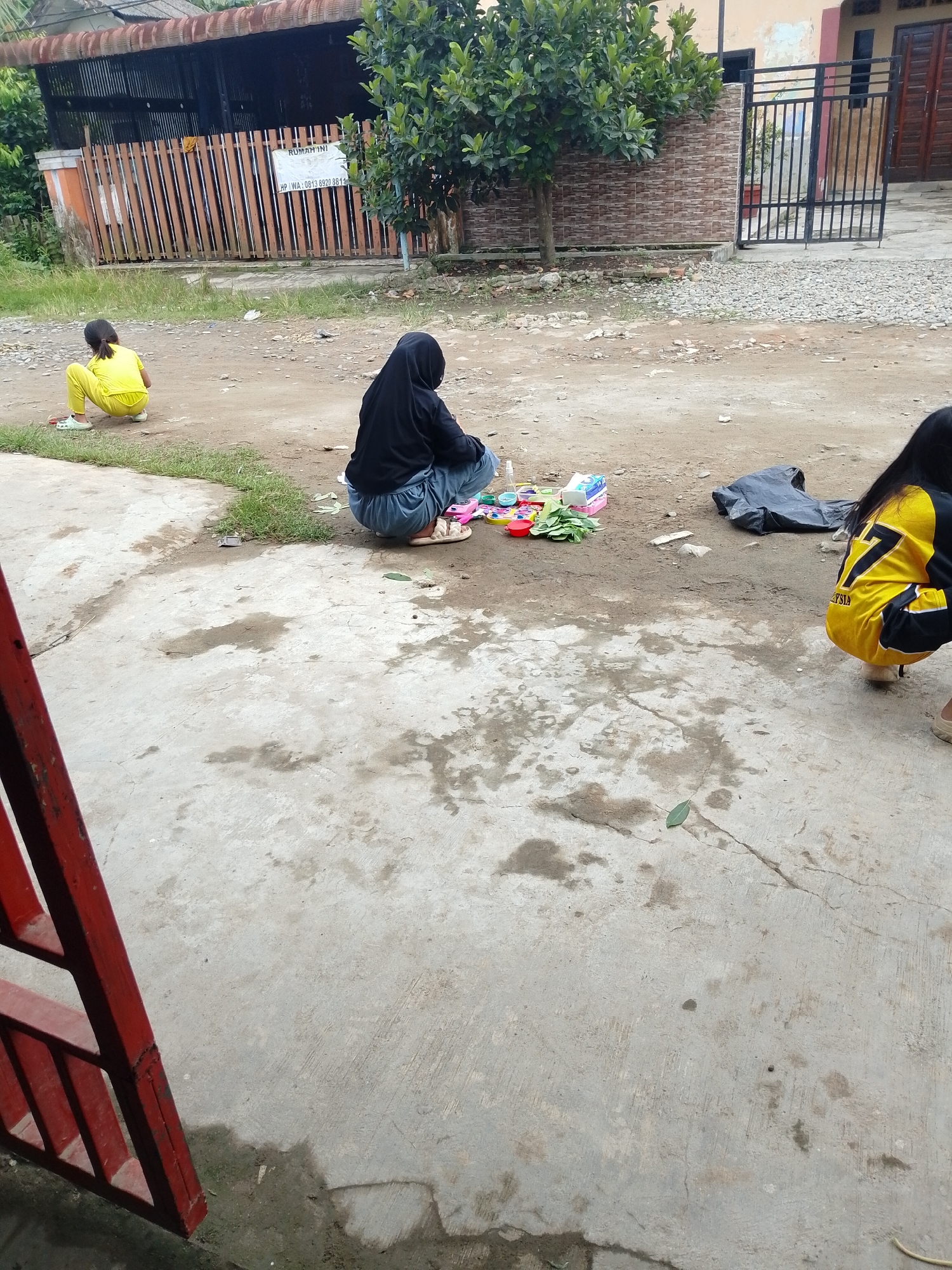 In Hamparan Perak, Indonesia, on a dirt and concrete ground, three young individuals are engaged in activities. To the left, a person wearing a yellow outfit crouches on the dirt, facing away from the camera. In the center, another individual, covered in a black hijab and light blue pants, also crouches, surrounded by various small objects including toy-like items, a tissue box, and green leafy material on the dirt. To the right, a third person in a yellow and black top with the number "7" on the back is crouched on the concrete, facing away from the viewer. The environment includes a wooden fence, a building with a tiled roof, and a metal gate in the background. The ground surface is uneven, with patches of dirt, gravel, and cracked concrete showing signs of wear and moisture.