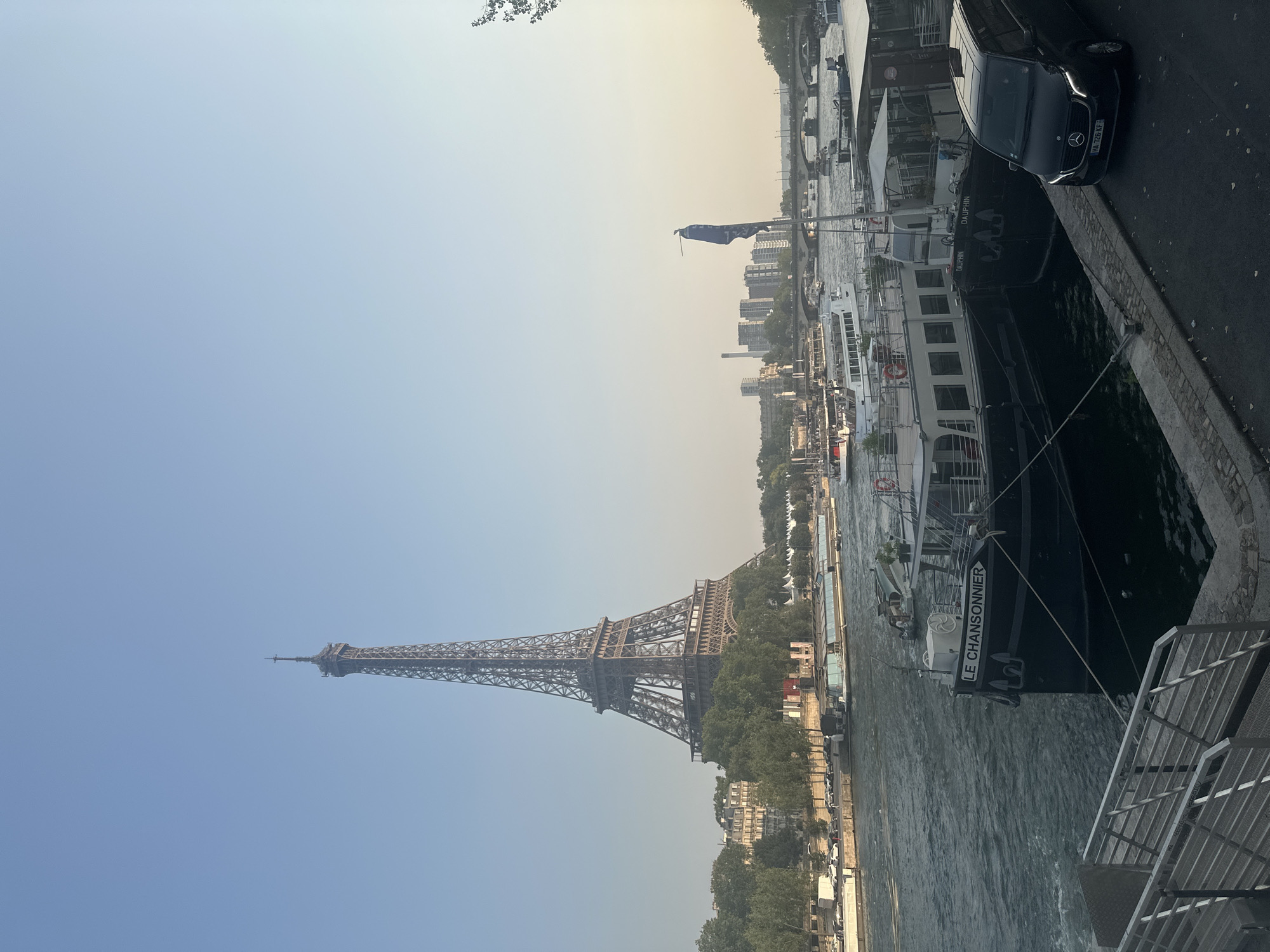 The image depicts a view of the Eiffel Tower in Paris, France, from across the Seine River. The Eiffel Tower is centrally located in the background, with the river flowing in the foreground. Several boats are docked along the riverbank, including a tour boat with the text "LE CHANSONNIER" visible on its side. A black van with a Mercedes-Benz logo is parked on the right side of the image, near the docked boats. The sky is clear and blue with a slight haze. The environment appears to be a public space along the river, with a walkway and railings in the foreground.