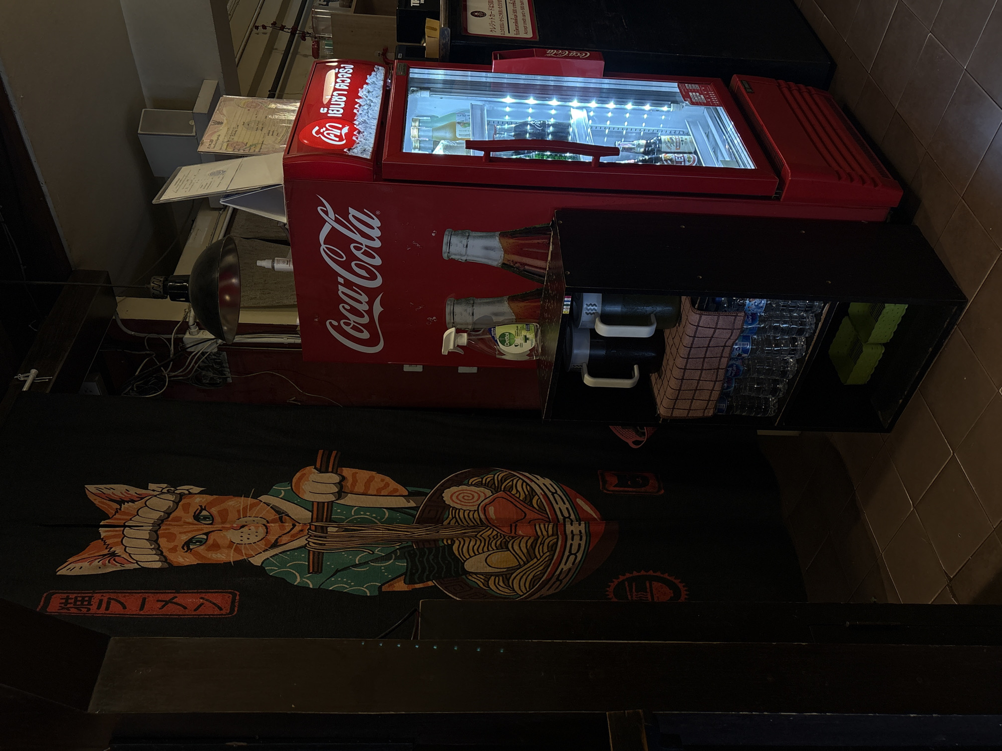 This image shows a Coca-Cola branded refrigerator filled with drinks, including bottles of Coca-Cola and other beverages. The refrigerator is red and features the iconic Coca-Cola logo. Next to it, there's a black cabinet with glasses and bottles. Below the refrigerator, a black tapestry with a Japanese-style illustration of a cat wearing a chef's hat and preparing ramen is visible. The tapestry also has Japanese text. Some signs with text in Thai and English are also present, indicating credit card payment policies. The overall scene appears to be in an establishment like a restaurant or bar, likely in Chiang Mai, Thailand, given the Thai text. The lighting suggests it could be indoors during the day or evening. There are no people visible in the frame.
