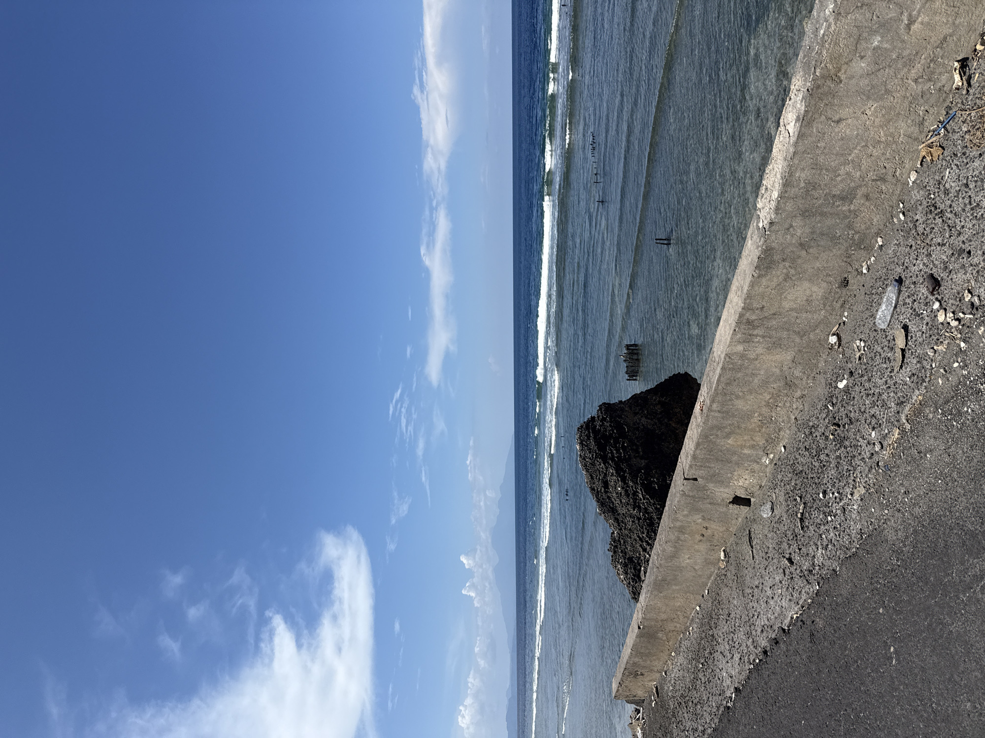 From Batununggal, Indonesia, the image shows a coastal scene. A concrete barrier and asphalt road are in the foreground. The sea extends towards the horizon, with small waves visible. A dark, rocky outcrop is positioned near the barrier. The sky is predominantly blue with scattered white clouds. Some wooden posts are located in the water.