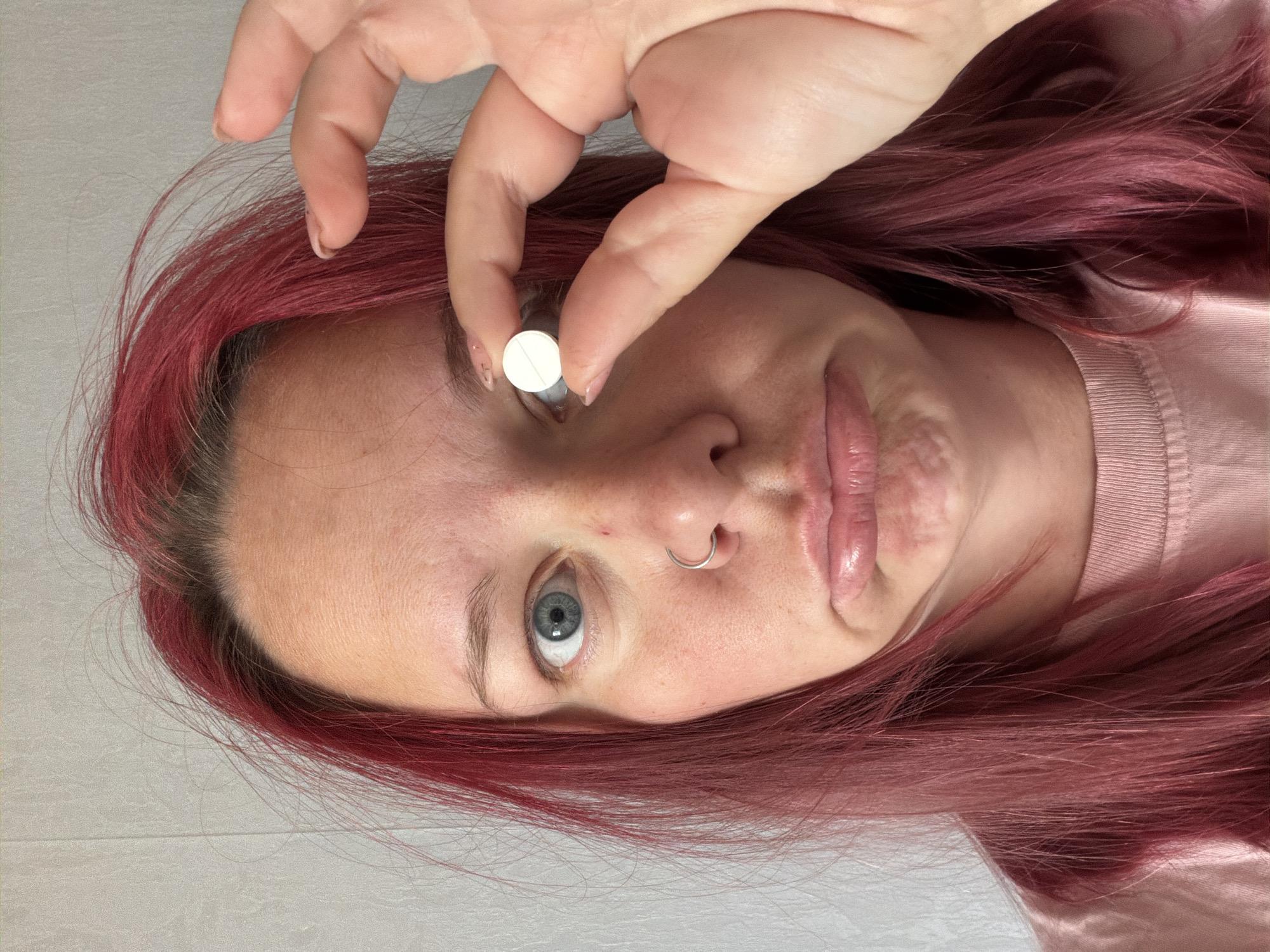 A person with dyed red hair is holding a small, round, white pill between their thumb and forefinger, positioned near their eyebrow. The person has blue eyes and a nose ring. The background is a light, solid color. The image is taken from a close-up perspective. There is no information in the image to suggest the location is Chaikovsky, Russia.
