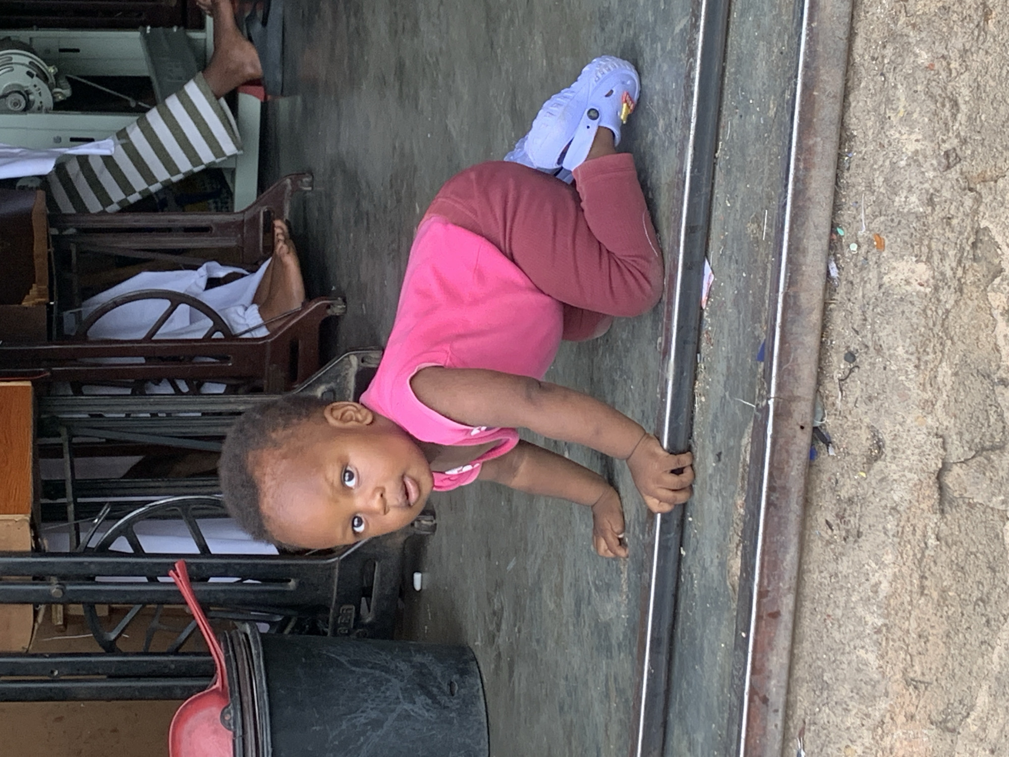 A toddler, wearing a pink shirt, maroon pants, and a light blue Croc shoe on one foot, is crawling on a gray surface. The child's head is turned towards the camera. To the left is a black sewing machine and a maroon dustpan. In the background, there is a person wearing striped pants sitting on a chair. The ground is concrete and textured. The photo was taken in Jos, Nigeria.