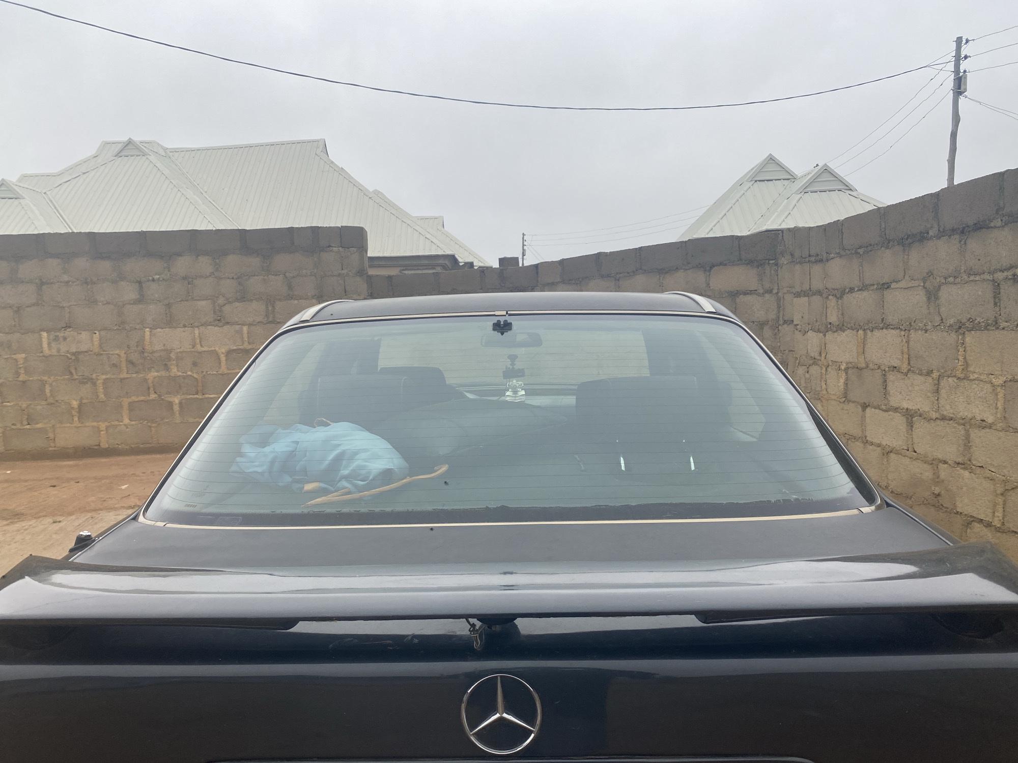 In Jos, Nigeria, a black Mercedes-Benz sedan is parked in front of a brick wall under an overcast sky. The rear of the car, including the trunk and taillights, is visible. A spoiler is attached to the trunk. Inside the car, a blue cloth is visible through the rear windshield, along with what appears to be a rearview mirror with a hanging ornament. The car is situated in what appears to be an outdoor residential area, with the roofs of two houses with white, metallic roofing visible behind the brick wall. The overall scene suggests a typical day in Jos, with the car being a central element in an everyday setting.