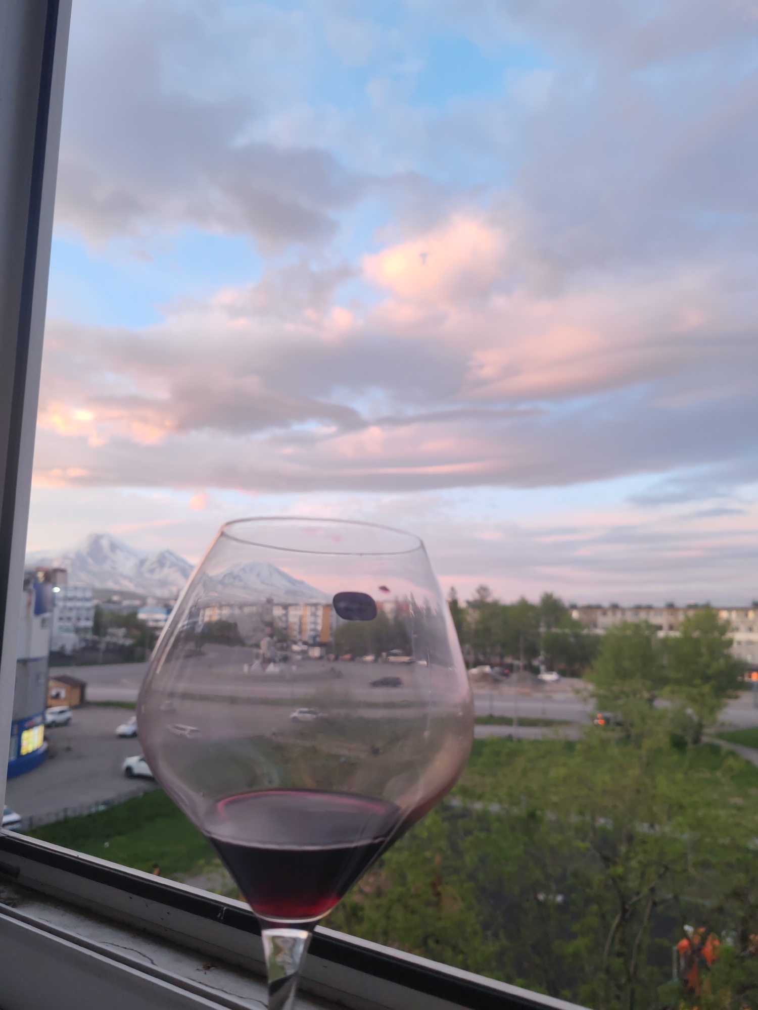 A glass of red wine is positioned on a windowsill in the foreground, with a cityscape and mountains in the background. The sky is a blend of blue, pink, and grey clouds, indicative of a sunset or sunrise. The cityscape appears to be Petropavlovsk-Kamchatsky, Russia, with apartment buildings and a street visible. Distant snow-capped mountains can be seen, likely the Vilyuchinsky volcano. The overall atmosphere suggests a moment of quiet contemplation or enjoyment of the view.