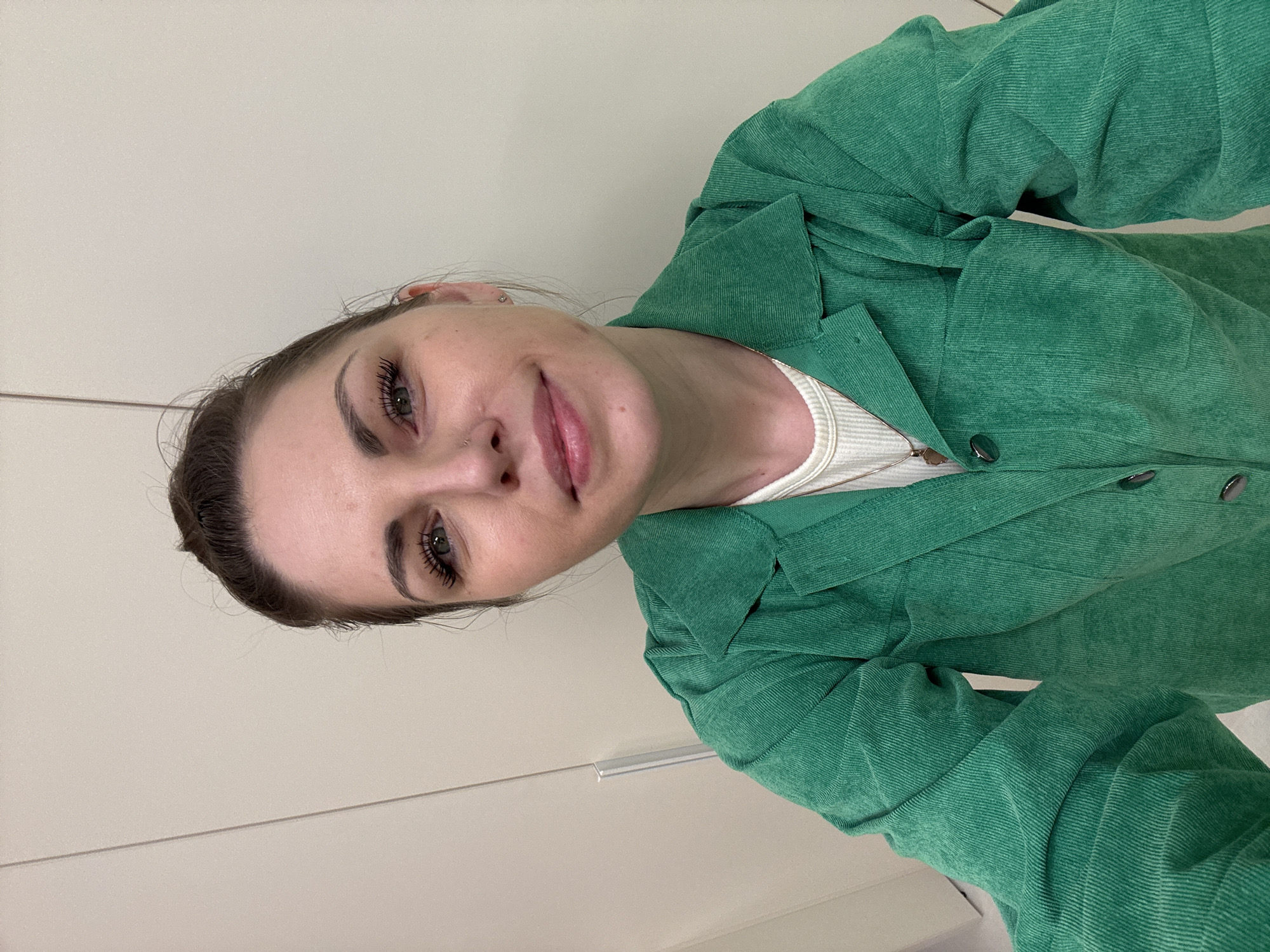 A young woman is depicted in a selfie, looking directly at the camera with a soft, closed-lip smile. Her head is tilted, conveying a relaxed and candid demeanor. She has dark hair pulled back, highlighting her face, which features well-defined eyebrows, prominent eyelashes enhanced with mascara, and eyeliner. A small metallic nose piercing adorns her left nostril.
She is wearing a dark green corduroy jacket or overshirt with visible buttons, layered over a light-colored, ribbed top. A slender gold-toned chain necklace is visible around her neck. The distinct ridged texture of the corduroy fabric is evident on her outerwear.
The background is a simple, light-toned wall or cabinet, featuring a subtle horizontal line that might be a seam or a drawer handle. The lighting is bright and uniform, suggesting an indoor environment. The image is taken at an angle, giving it a dynamic perspective. There is no visible text in the media. This scene takes place in Klin, Russia.