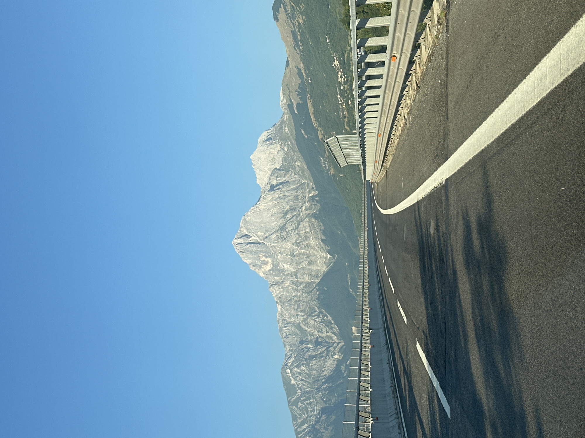 The image shows a highway in Ornano Grande, Italy, with a large mountain range in the background. The highway has two lanes with white lane markings and a concrete barrier with a metal guardrail on the right side. The sky is clear and blue. The mountain range is rocky and barren at the top, with green trees covering the lower slopes. A small village is visible on the hillside in the distance. The image appears to be taken from a moving vehicle, as indicated by the perspective and the shadows on the road.