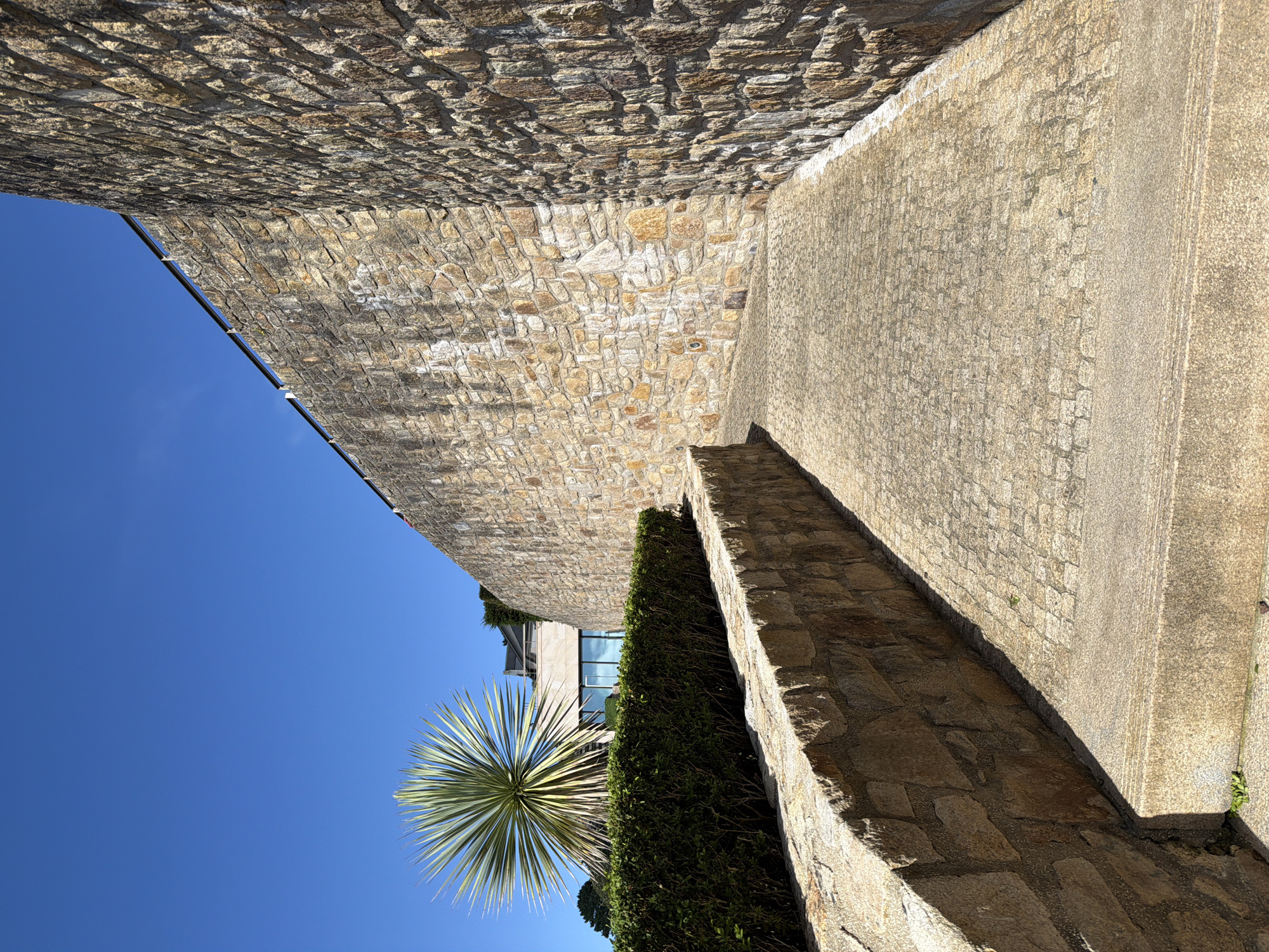 This media file captures an outdoor scene with a strong architectural presence, bathed in bright sunlight. The primary subjects are two contrasting walls, one constructed of rough, uneven stone blocks and the other a smoother, textured concrete. These walls create a narrow pathway or corridor, leading the viewer's eye into the scene.
To the left, a large expanse of clear, vibrant blue sky dominates, suggesting a sunny day. A large, spiky plant, possibly a yucca or agave, is visible against this sky, its green leaves fanning out dramatically. Behind the plant, a modern building with large glass windows can be seen, partially obscured by dense green hedging.
The pathway itself is paved with irregular stone slabs. The stone walls are a warm, earthy tone, with visible mortar between the individual blocks. The concrete wall is lighter in color and has a more uniform, granular texture. Shadows are cast by the walls and the plant, indicating the sun is high in the sky, likely mid-morning or afternoon.
The overall impression is one of a well-maintained outdoor space, possibly part of a private residence, a hotel, or a public garden. The combination of natural elements like the plant and stone with modern architecture suggests a contemporary design. The lighting and clear sky convey a sense of warmth and pleasant weather. There is no visible text in the image. The location context provided is La Clarté, France, which aligns with the Mediterranean-like vegetation and stonework.