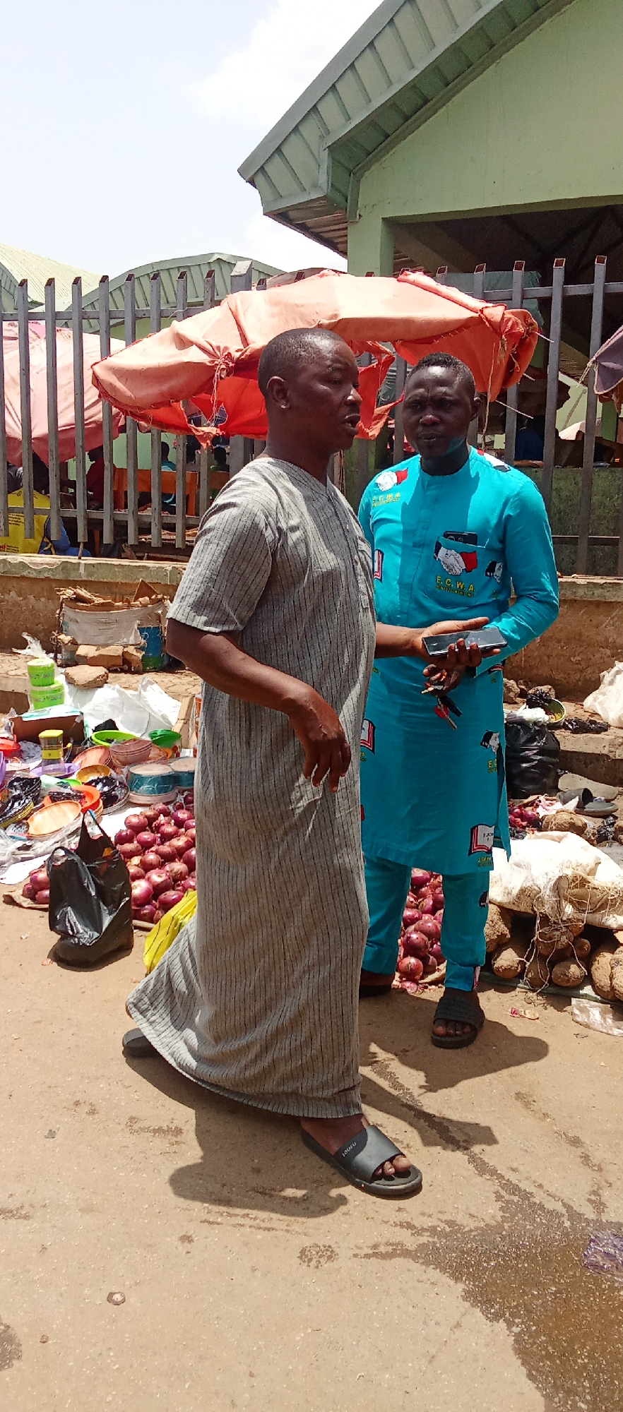 The image shows two men standing in a marketplace in Akwanga, Nigeria. The first man is wearing a long, gray striped robe and dark sandals. The second man is wearing a bright blue, long-sleeved tunic and matching pants with an "ECWA" emblem and black sandals. He is holding a black smartphone. In the background, there is a stall with produce, including onions, yams, and other goods. A faded red awning covers part of the stall. A black plastic bag sits on the stall. A metal fence is also visible in the background. The setting is a sunny day with some shadows.