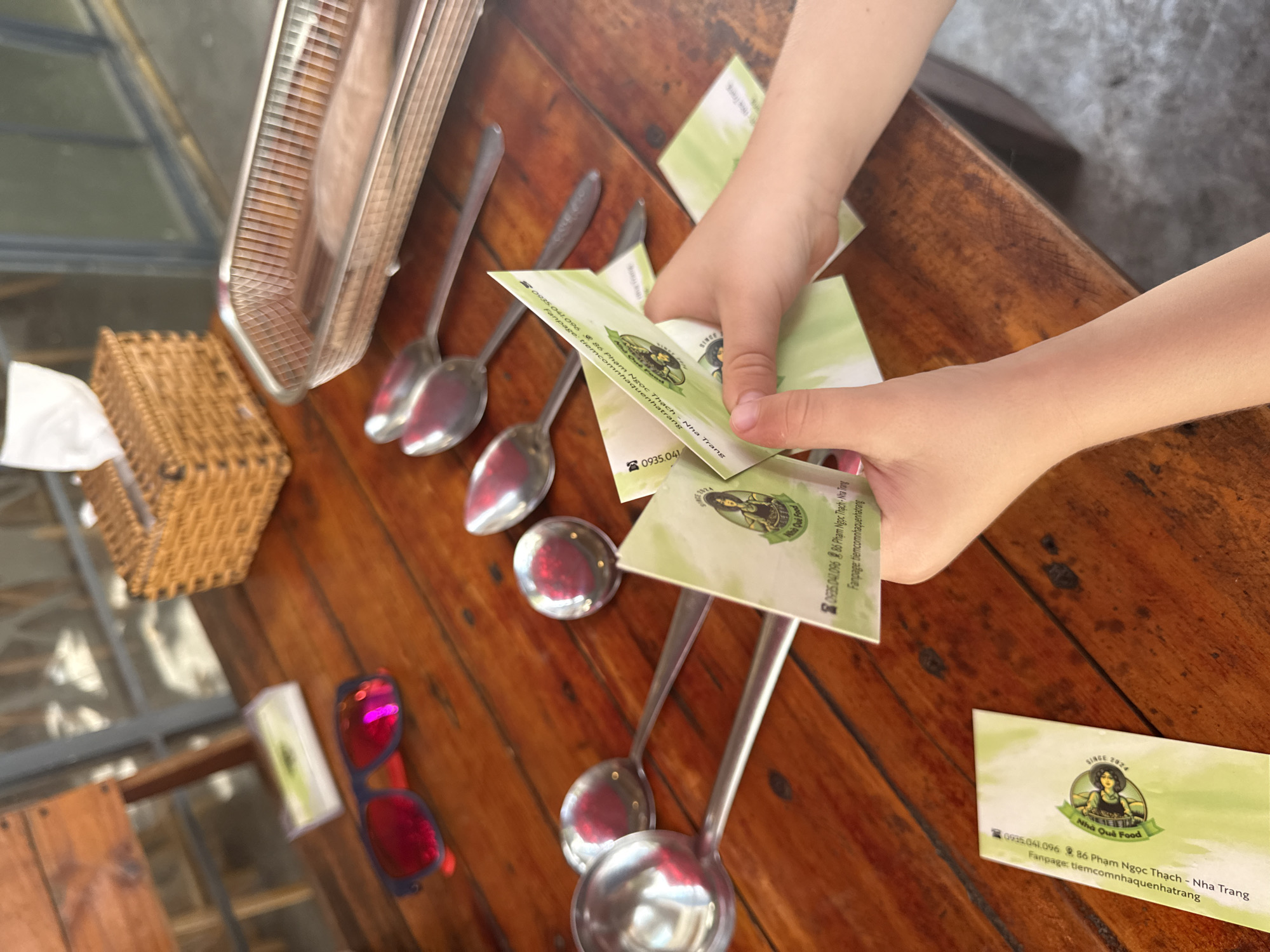 The image captures a close-up view of a wooden table with various objects. In the foreground, a person's hands are seen holding and manipulating several business cards that appear to be from a Vietnamese establishment named "Nha Qui Food." The cards have text in Vietnamese and a logo featuring a woman. The text on the cards indicates an address at "86 Phạm Ngọc Thạch - Nha Trang" and a Facebook page "fanpage:tiemcomnhaquienhatrang."
Scattered across the table are several large metal spoons and ladles, some reflecting the red of sunglasses also present on the table. To the right, a metal tray holds what appears to be some food item. In the background, a wicker tissue box sits on the table. The environment suggests an outdoor setting with a wooden chair and railing visible. The overall scene is casual, possibly a meal or a visit to a food establishment in Nha Trang, Vietnam.