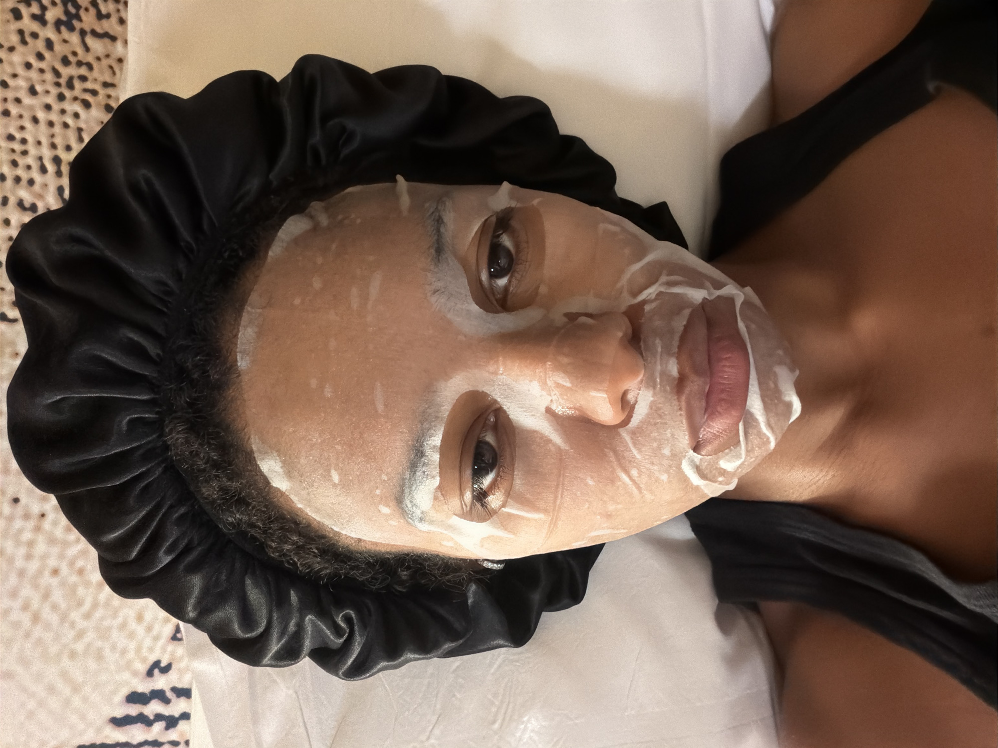 A close-up, eye-level shot captures a person lying down with a white sheet mask on their face. They are wearing a black satin bonnet that covers their curly hair. The mask covers most of their face, with openings for the eyes, nose, and mouth. White foam from the mask is visible around the edges and on the lips. The person is wearing a black tank top. The background consists of white bedding and a textured surface, possibly a headboard or wall covering. The scene appears to be indoors, in a well-lit room. There is no indication of the specific location within the city of Bremen, Germany.
