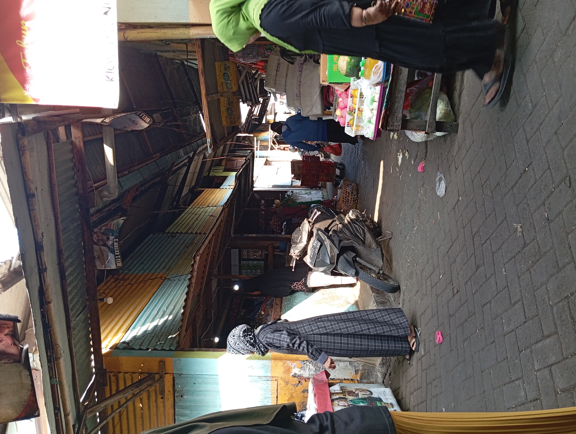 The image depicts a bustling outdoor market in Kepulungan, Indonesia. In the foreground, a woman wearing a green hijab and a long black dress walks towards the right, partially obscuring a fruit and vegetable stall. The stall displays a variety of produce, including watermelons, apples, and bottled drinks. To the left, a woman in a grey checkered dress and headscarf stands facing a stall. A motorcycle is parked on the paved ground between the two women. The market stalls are constructed with corrugated metal and wood, with awnings providing shade. The background shows more market stalls and activity. Sunlight filters through gaps in the overhead structures, casting shadows on the ground. The overall scene captures the vibrant atmosphere of a local Indonesian market.