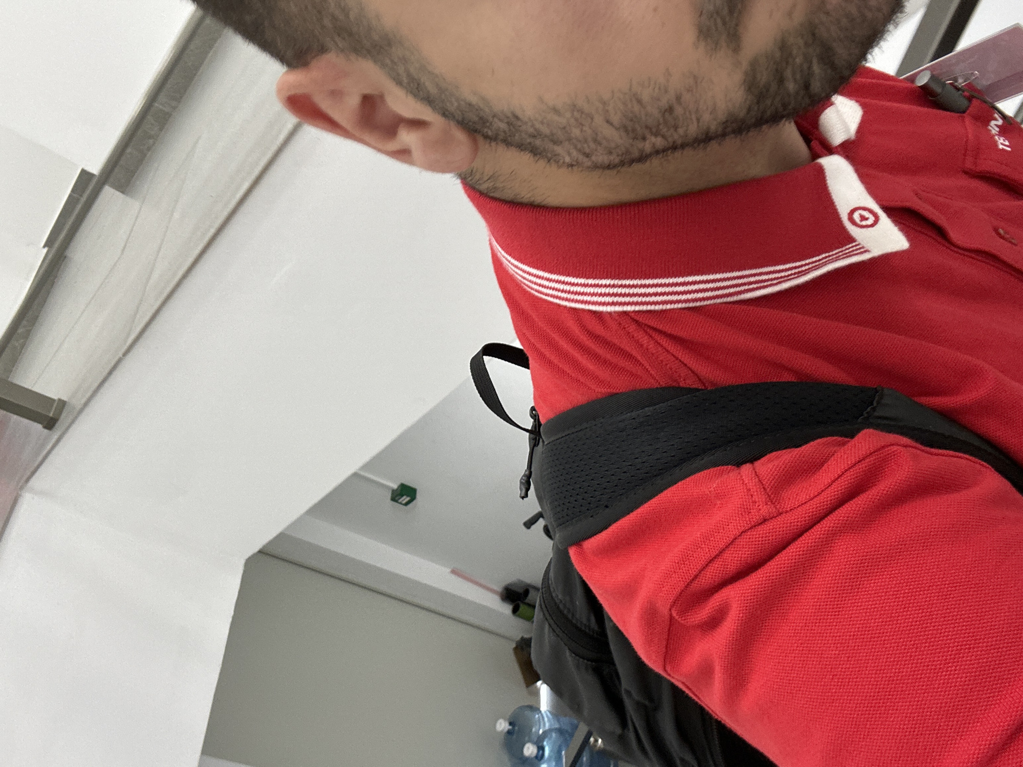 The image shows a person with a beard, wearing a red polo shirt with a white striped collar, and a black backpack. The person is facing slightly away from the camera. The background consists of a white wall and ceiling. The location is Lovech, Bulgaria.