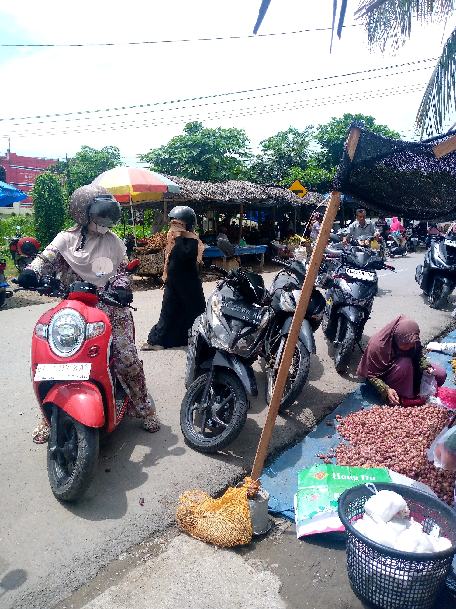 The image depicts a bustling street market scene in Keudeteupinpunti, Indonesia, under an overcast sky. In the foreground, a red Honda scooter is parked, ridden by a person wearing a beige hijab and a patterned dress, and a helmet. To the right of the scooter, a woman in a black hijab is standing, appearing to interact with a vendor. Further to the right, a woman wearing a maroon hijab is kneeling by a large pile of small, red onions spread out on a blue tarp, and is bagging some of them. Several other motorcycles are parked around the market. In the background, there are market stalls with thatched roofs, displaying various goods, and more people are visible browsing or walking through the market. Trees and buildings can be seen beyond the market stalls. A yellow traffic sign is also visible. The ground is paved, with some debris and a small puddle of water.