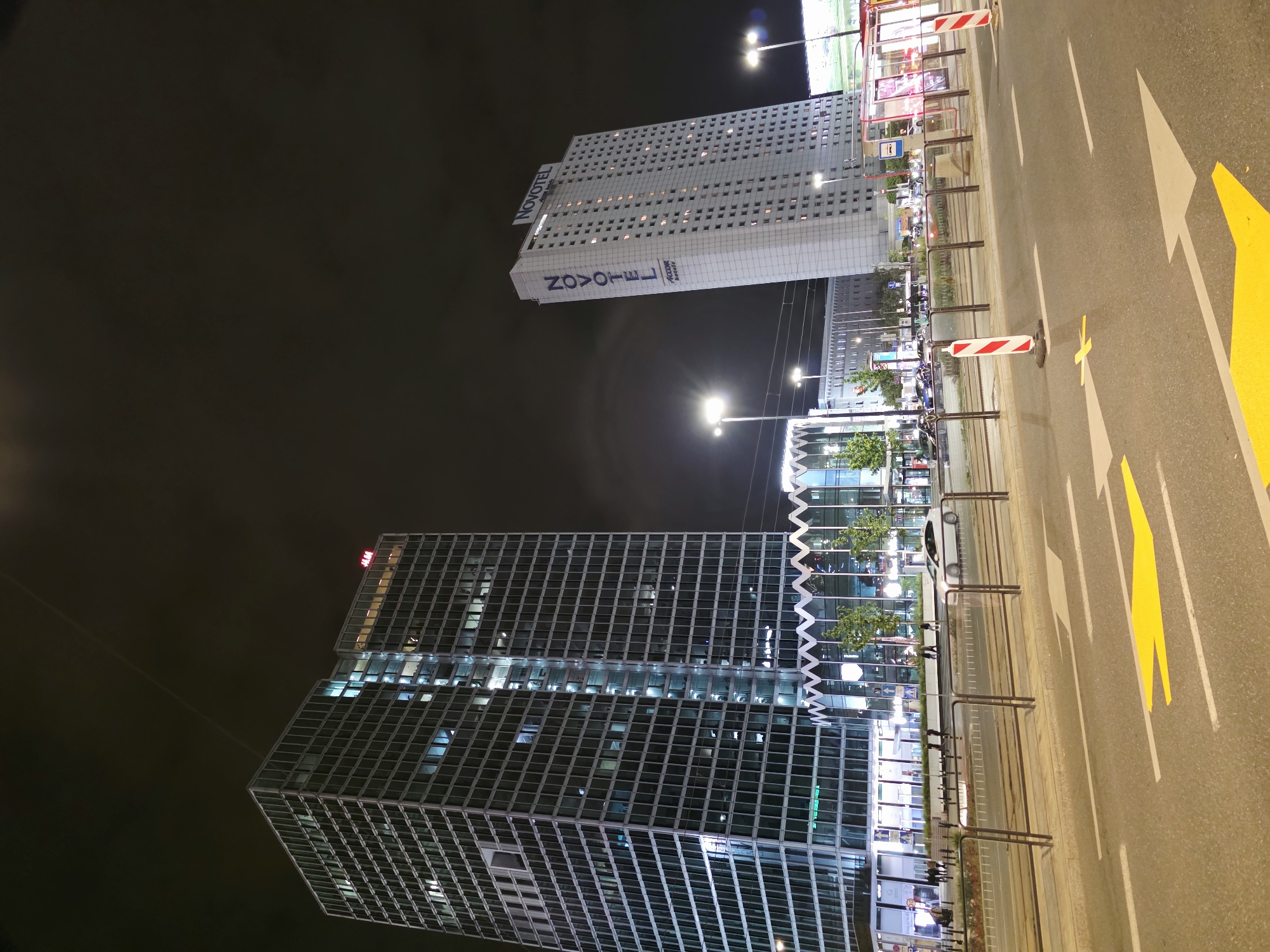 This is a night-time, low-angle, wide shot of the intersection of Towarowa and Jerozolimskie avenues in Warsaw, Poland. Two prominent buildings dominate the scene: a modern glass skyscraper on the left, identified by the "H&M" logo at its top, and the Novotel hotel building on the right, featuring its name vertically on its facade. The foreground shows a street with lane markings, including yellow arrows, indicating traffic flow. A tram stop with illuminated advertisements is visible on the right side of the image. Streetlights illuminate the scene, casting a glow on the dark sky and the buildings. The street is relatively clear, with a few vehicles and pedestrians visible in the middle ground. The architecture suggests a modern urban environment with a mix of commercial and hospitality establishments.