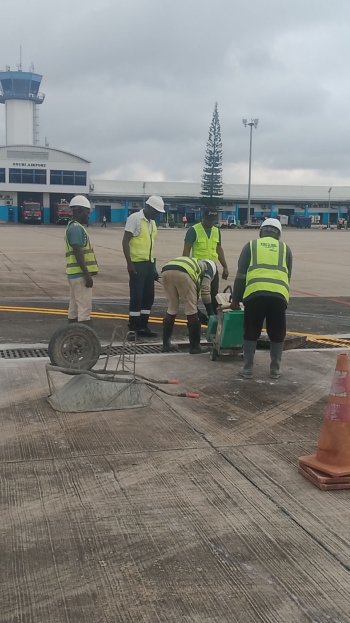 The image depicts a scene at Osubi Airport in Orerokpe, Nigeria. Four workers in high-visibility vests and hard hats are gathered around a piece of machinery on a concrete surface. One worker appears to be operating the machine. A wheelbarrow lies tipped over on the ground nearby, and an orange traffic cone sits to the right. In the background, the airport control tower is visible, as is the airport building with a tall pine tree nearby. There are emergency vehicles parked near the airport building. The sky is overcast.