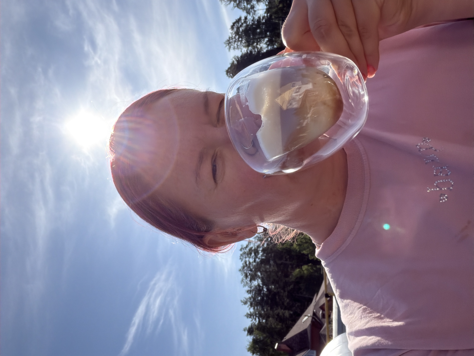 A woman with pink hair is holding a clear glass up to her face, presumably to drink from it. The glass contains a liquid that appears frothy and white. The background consists of a bright blue sky with some clouds and a few trees. There appears to be a house in the background as well. The location is not identifiable as Chaikovsky, Russia, based on the content of the image. The woman is wearing a pink shirt with some kind of reflective embellishment on it.