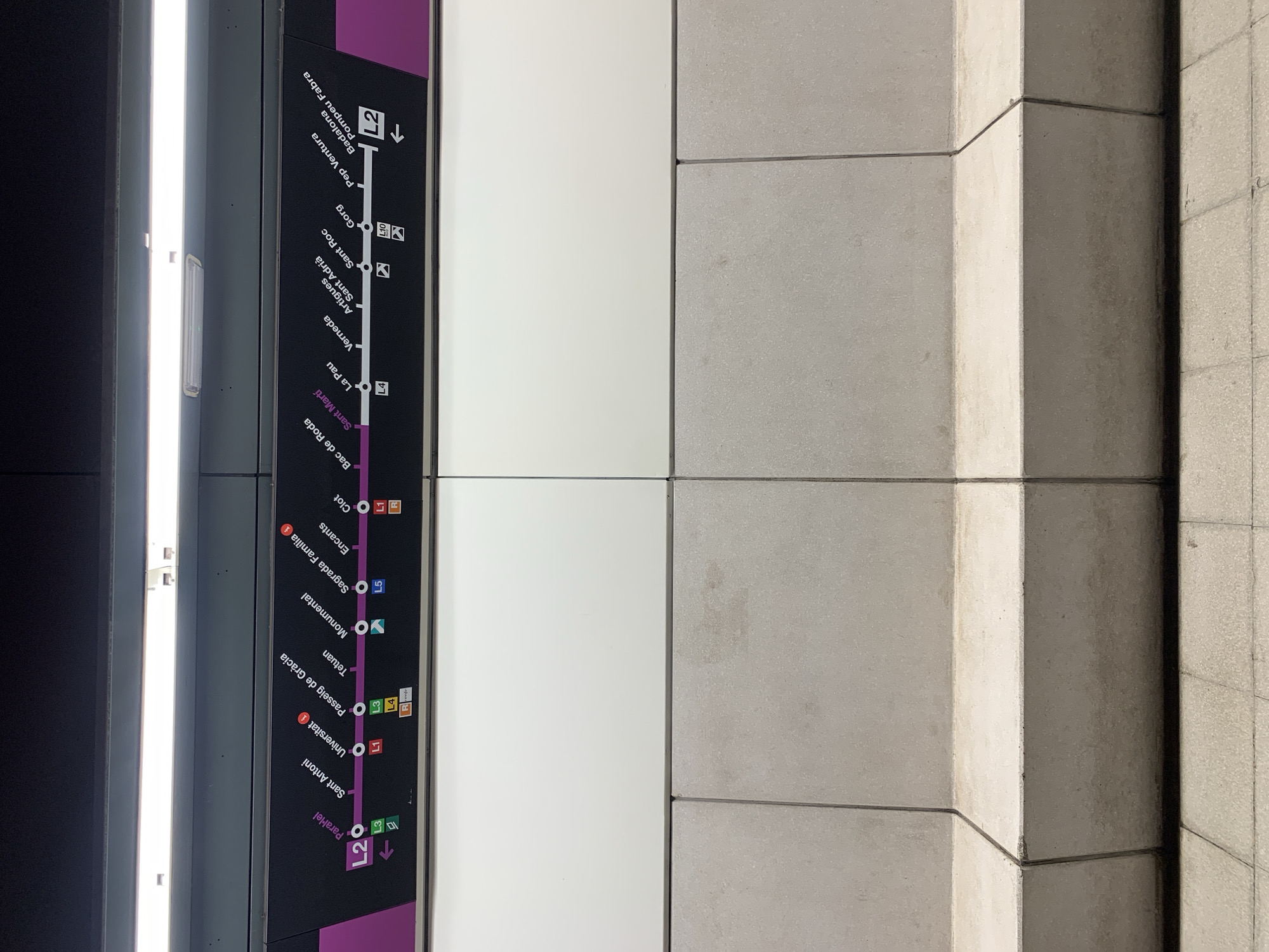 The image captures a static view inside what appears to be a metro station in Barcelona, Spain. The primary subject is a dark-colored information panel displaying a section of a metro line map, positioned vertically along the left-center of the frame. To its immediate left is a tall, bright fluorescent light fixture, casting a strong glow. To the right of the map panel, a light grey, tiled wall with distinct grout lines forms the backdrop, characterized by its textured, concrete-like surface and a prominent corner or pillar creating a shadow.
The metro map prominently features Line 2 (L2), indicated by a thick purple line. Numerous station names are clearly visible along this line, indicating connections to other metro lines (e.g., L1, L3, L4, L5, L10) and Renfe commuter trains with corresponding color-coded icons. Stations listed include "Paral·lel," "Sant Antoni," "Universitat," "Passeig de Gràcia," "Tetuan," "Monumental," "Sagrada Família," "Encants," "Clot," "Bac de Roda," "Sant Martí," "La Pau," "Verneda," "Artigues Sant Adrià," "Sant Roc," "Gorg," "Pep Ventura," and "Badalona Pompeu Fabra." Small arrow icons on the map denote the direction of travel and transfer points.
No people are visible, and there is no discernible activity taking place. The scene suggests an operational public transport environment. The lighting conditions are consistent with an indoor setting, and no specific time of day or weather can be inferred. The overall impression is one of functionality and utility, common in urban transit infrastructure.