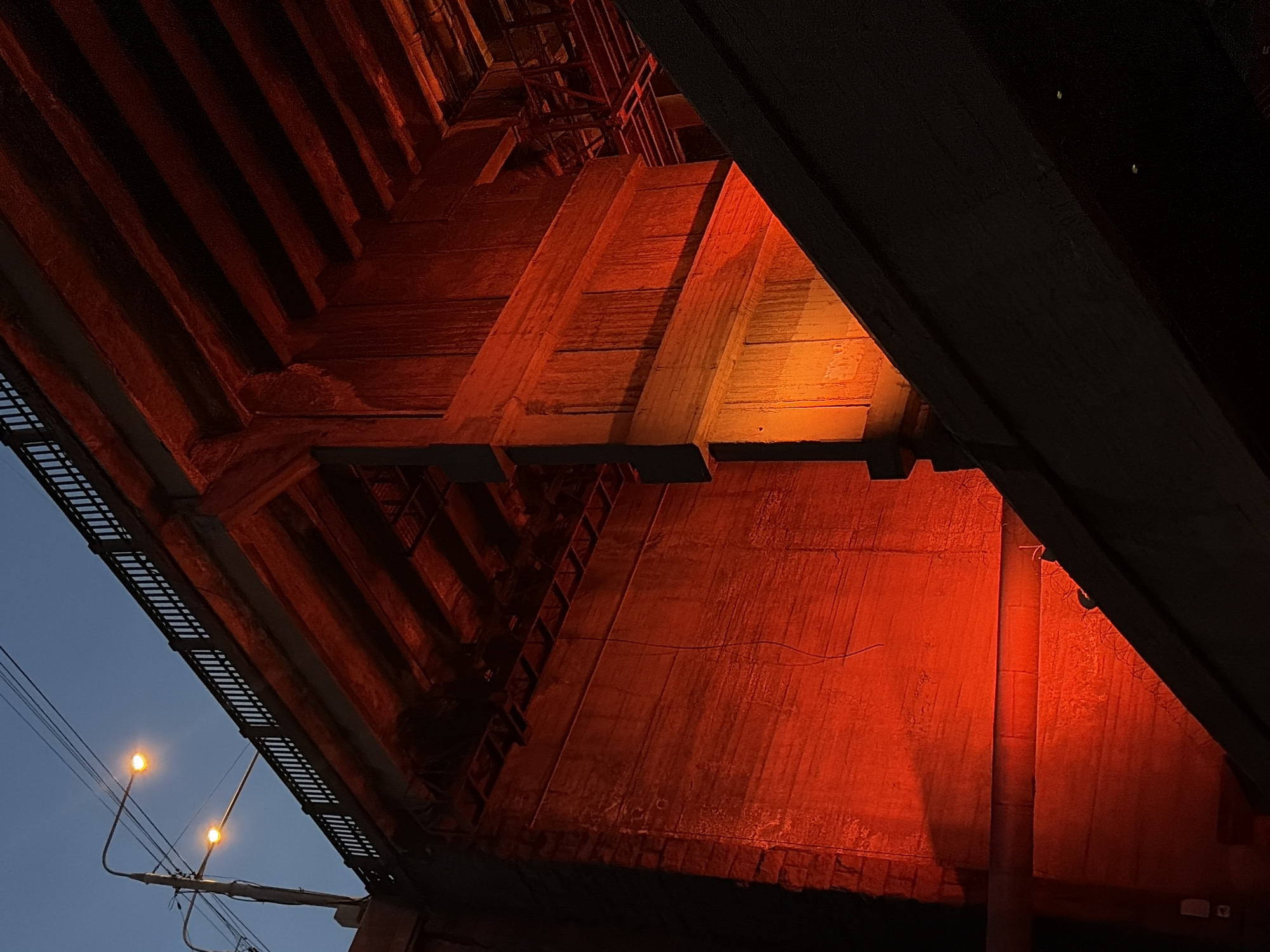 The image depicts the underside of a bridge in Krasnoyarsk, Russia, illuminated by red and orange lights. The bridge structure, made of concrete and metal, is visible with its supporting beams and railings. Two streetlights are also visible in the upper left portion of the frame against a dark blue sky. The scene is devoid of any people or discernible activity. The lighting creates a dramatic and somewhat stark atmosphere.