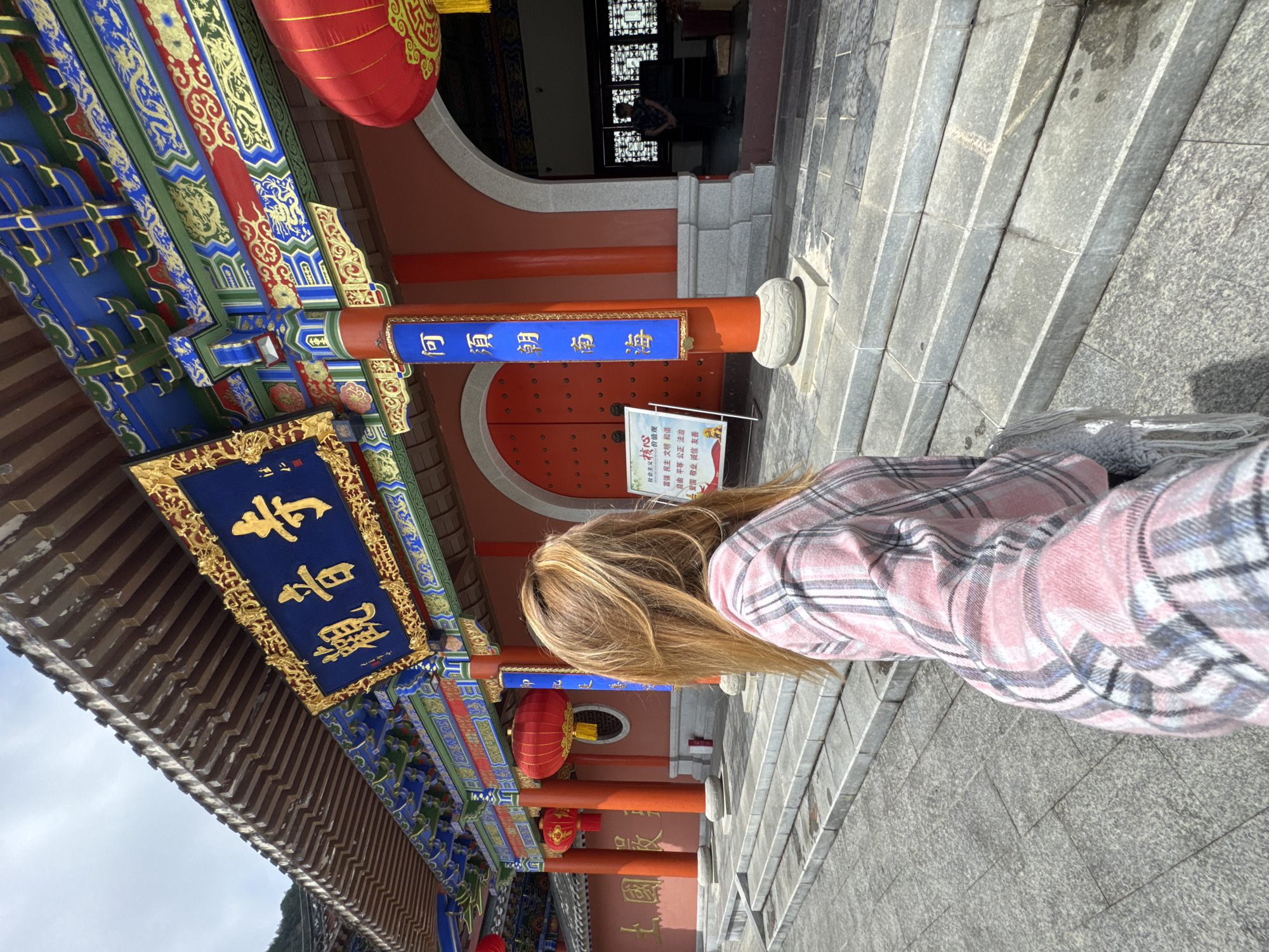 The image shows a woman with blonde hair, wearing a pink and gray plaid shirt, standing on a stone staircase in front of a traditional Chinese building. The building has ornate decorations with vibrant colors like blue, red, and gold. Signs with Chinese characters are visible, including "觀音寺" (Guanyin Temple) and phrases related to socialist core values and the inscription "何須朝南海" (Why bother worshiping towards the Southern Sea). Red lanterns hang from the roof. The scene appears to be in Qianjiang, China, indicated by the Chinese signage and architectural style.