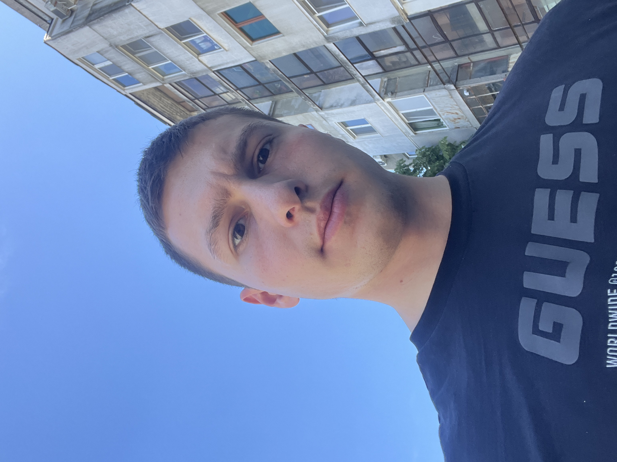 A man stands beneath a building in Burgas, Bulgaria. The man is wearing a dark blue GUESS t-shirt. The sky is bright blue. The building behind the man has many windows.