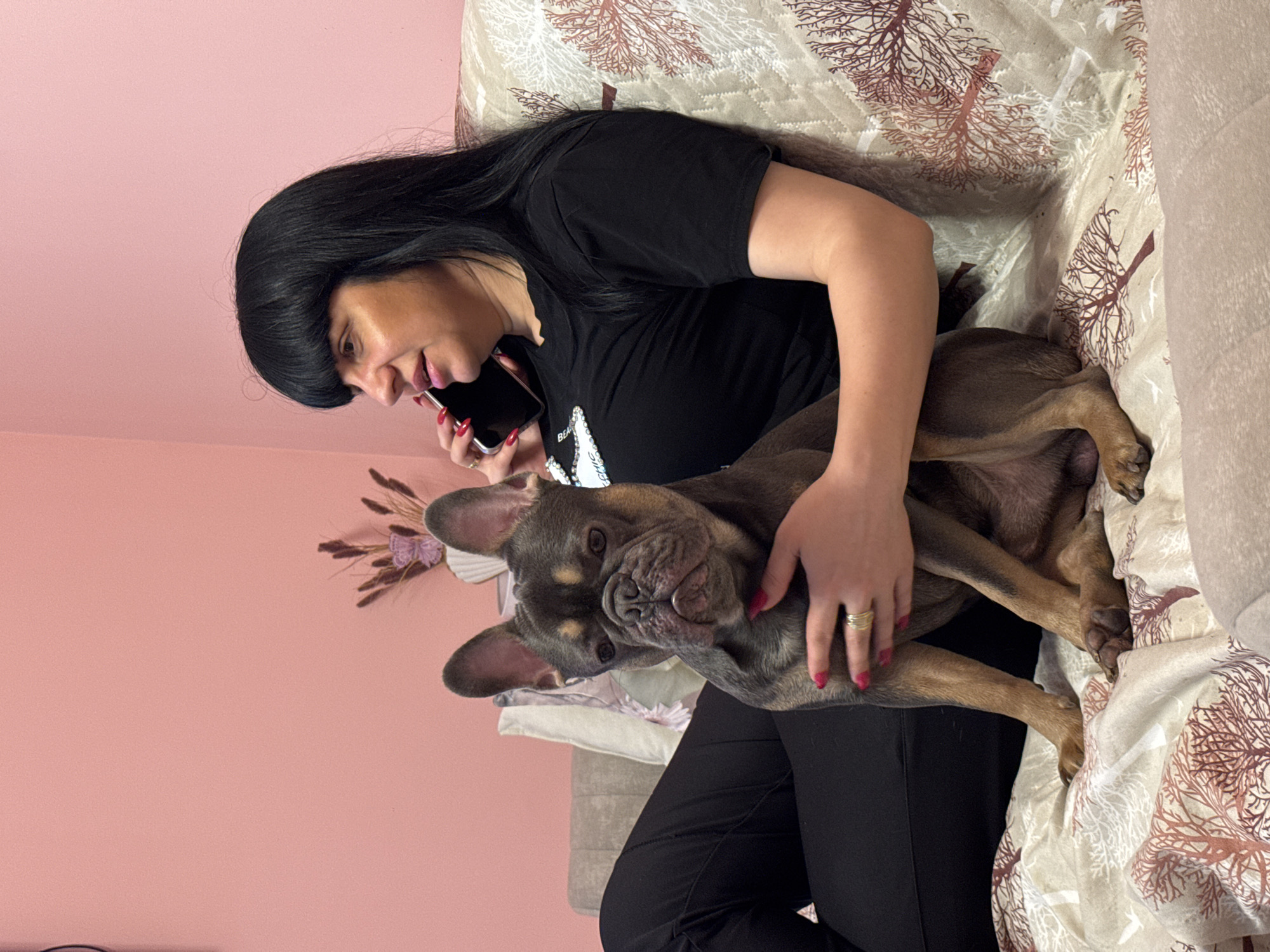 A woman with dark hair is sitting on a patterned couch, holding a French bulldog and talking on a smartphone. The scene is set indoors against a light pink wall decorated with dried plants in the upper left corner. The woman is wearing a black t-shirt and black pants. She has pink nail polish. The couch is covered in a patterned fabric with brown tree-like designs. The dog is a gray/brown color. The location is possibly a home in Burgas, Bulgaria.
