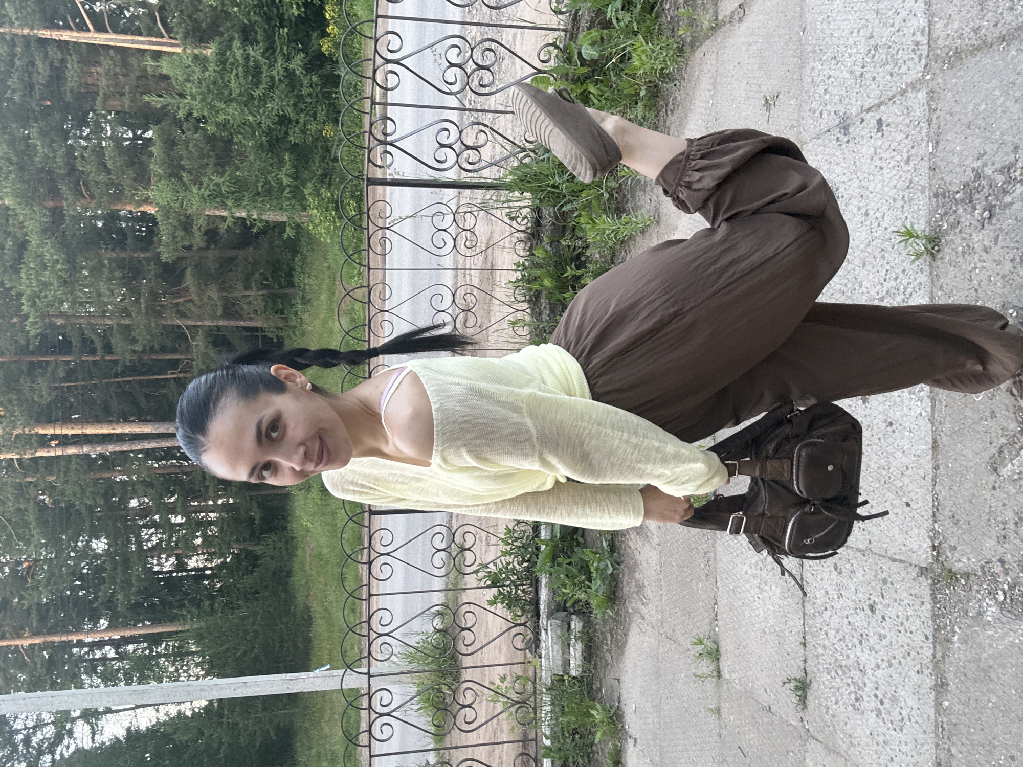 This image captures a female individual posing outdoors on a concrete sidewalk in the city of Votkinsk, Russia. The subject, an adult with dark hair pulled back into a ponytail featuring a visible braid, is smiling faintly while looking directly at the camera. She is dressed in a light yellow, textured, loose-fitting long-sleeved top, worn off the right shoulder to expose a light pink bra strap. She wears flowy, wide-leg dark brown pants, with a ruched detail visible on the cuff of her left leg. Her left leg is bent and raised behind her, displaying the dark, ridged sole of her flat shoe. In her left hand, she carries a dark brown, unstructured shoulder bag with multiple zippered and flap pockets. The immediate foreground consists of a mottled concrete sidewalk with small weeds growing through cracks. Behind the subject, a black wrought-iron fence with heart and scroll motifs runs horizontally, separating the sidewalk from a grassy strip and an asphalt road. In the background, a dense coniferous forest, predominantly pine trees, stands under natural outdoor lighting. A light-colored utility pole is visible on the far left.