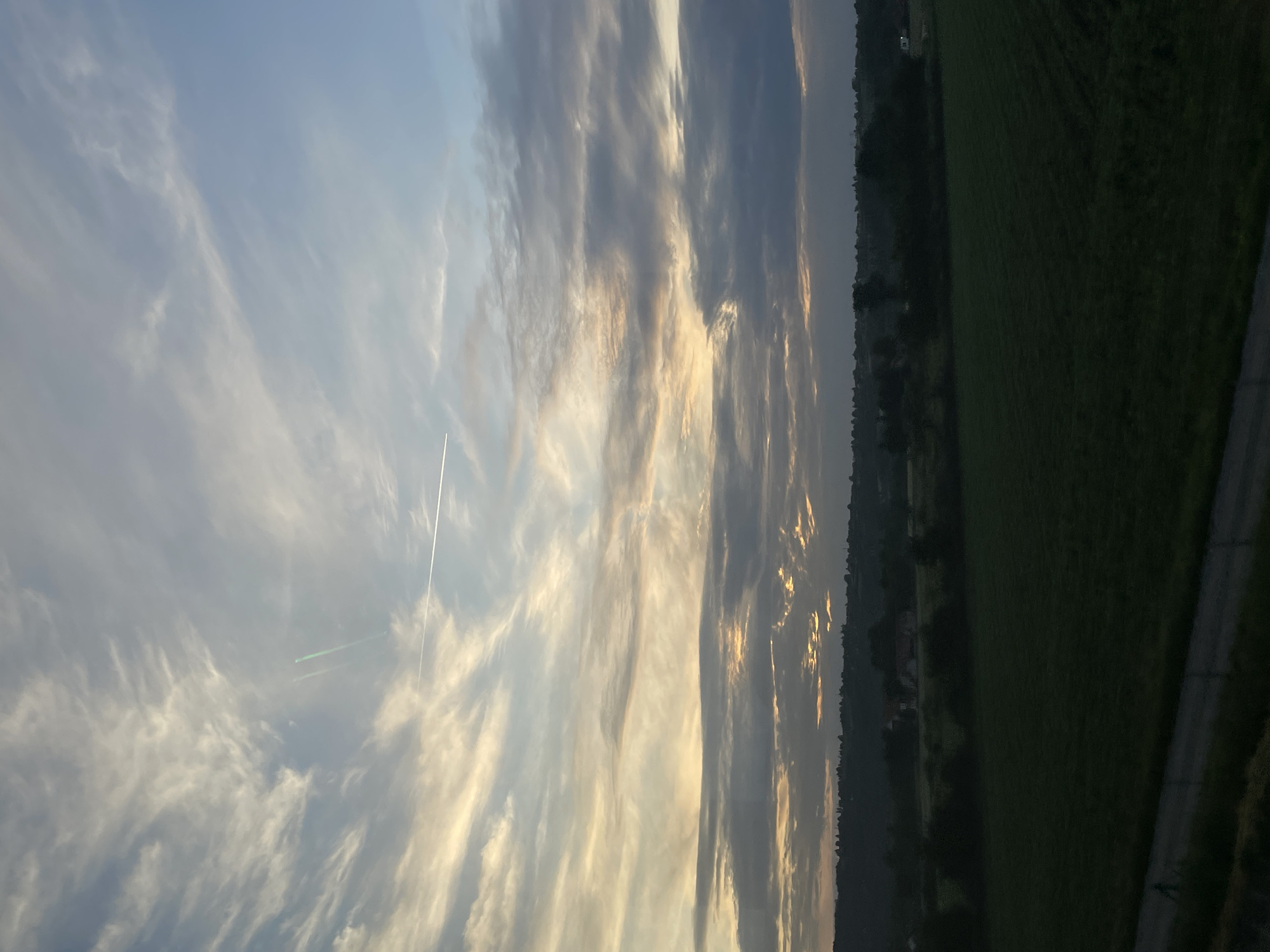 The image depicts a landscape at an indeterminate location in Obrenovac Urban Municipality, Serbia. Dominating the left half of the frame is a sky filled with feathery clouds illuminated by the setting sun. Contrails, possibly from an aircraft, are visible in the upper left quadrant. The right half of the image features a dark, tree-lined horizon bordering a large, green field. A road, partially visible in the lower right corner, runs alongside the field.