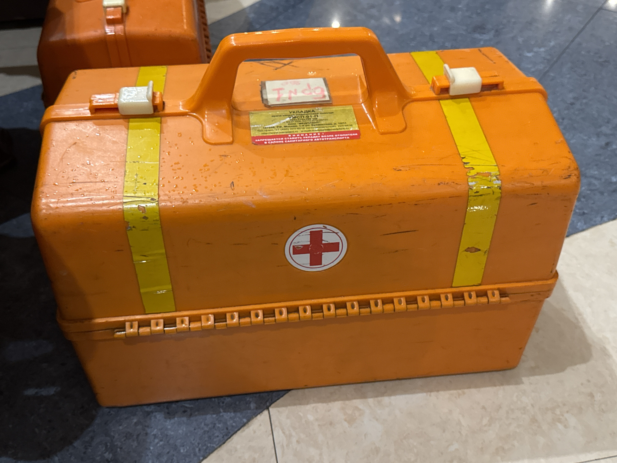 Here's a detailed description of the image:
**Subject:** The image features a bright orange medical kit. The kit has a handle on top, two white latch closures on either side of the handle, and yellow stripes running vertically down the sides. A red cross is centered on the front panel within a white circle. The lower edge of the top section has protruding loops. There are two labels on the top near the handle. One handwritten in ink and another printed.
**Environment:** The kit is placed on a surface consisting of light grey and tan tiles. A portion of a similar medical kit is visible in the upper left corner of the image.
**Notable Details:**
* The medical kit has a used and worn appearance, with scratches and scuffs visible on the surface.
* The printed label appears to contain information in Russian, possibly indicating the origin or intended use of the kit.
**Possible Location:** The location is unknown. The floor tiles and the medical kit itself don't provide enough context to determine the city or even indoor/outdoor setting definitively.