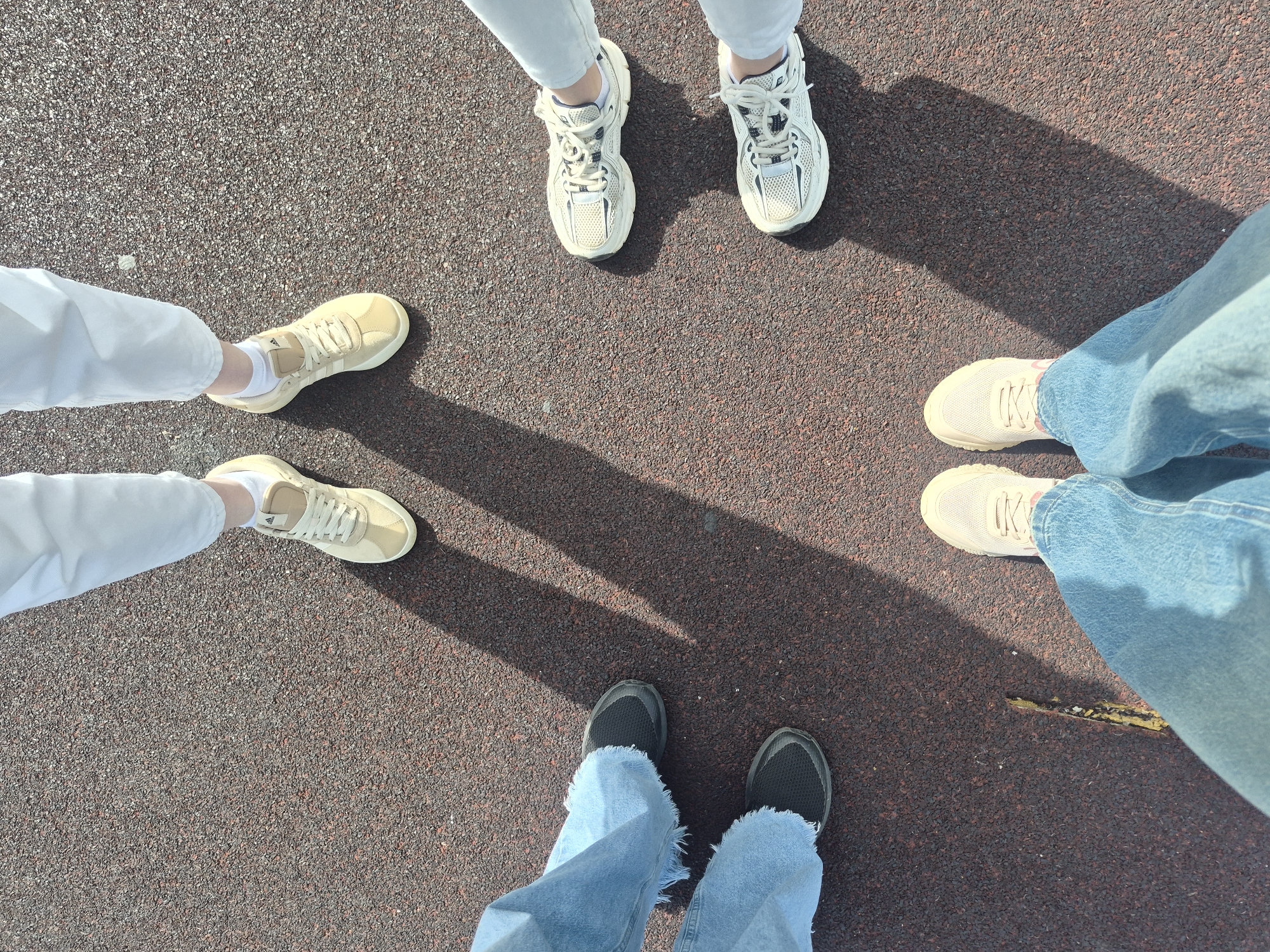 The image shows a ground-level view of five people standing in a circle. The focus is on their footwear and the lower portion of their legs. One person is wearing white athletic shoes with white pants. Two people are wearing tan shoes, one paired with white pants and the other with blue jeans. Another person is wearing beige sneakers paired with blue jeans. The final person is wearing black athletic shoes with frayed blue jeans. The ground surface appears to be a maroon-colored asphalt or paved surface. The lighting suggests it is daytime with some shadows cast on the ground. There are no other visible environmental details to ascertain a specific location.
