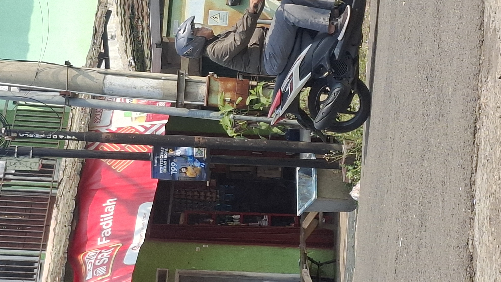 A person rides a motorcycle on a paved road in front of a building with a red banner. The banner reads "SRC Fadilah". A sign on a utility pole displays "WIFI TERCEPAT" and a price of "199,-". A green wall is visible behind the banner. A person is wearing a helmet and a jacket while riding the motorcycle. The environment appears to be a street scene in a town or city.
