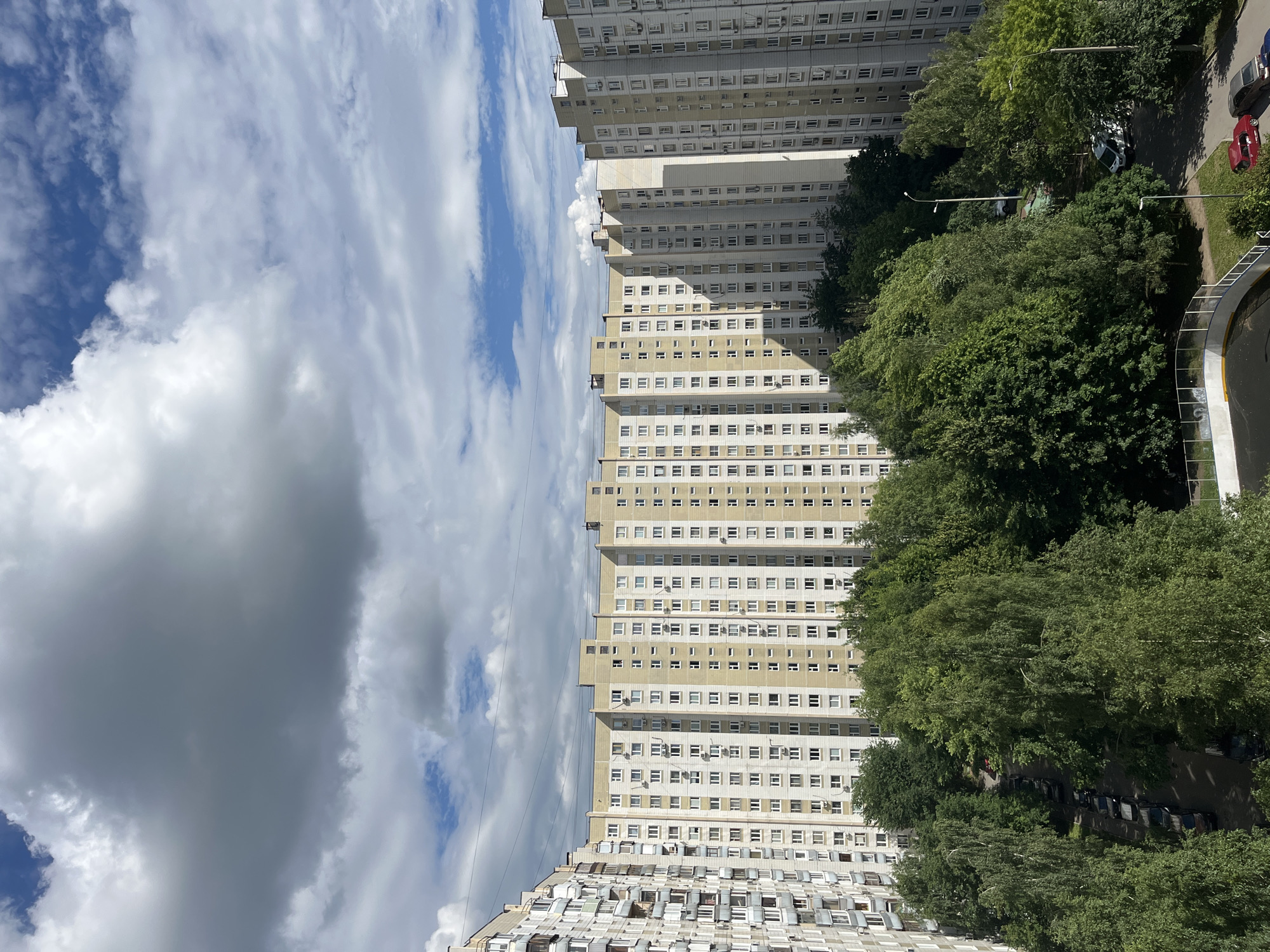 This media file depicts a view of apartment buildings and trees under a cloudy sky. The buildings are tall, multi-story structures, appearing to be residential. They are lined with numerous windows and balconies, some of which are enclosed with glass or have air conditioning units attached. One building in the foreground has a more complex facade with many smaller windows or extensions.
A dense cluster of green trees borders the apartment buildings, suggesting a park or a well-treed urban area. Sunlight filters through the clouds, casting shadows on the buildings and the trees, indicating it is daytime. The sky is filled with a mix of white and gray clouds, suggesting a partly cloudy or overcast day.
In the background, a street is visible with several cars parked along the side, including a red car and a darker colored vehicle. There is also a white car on the street. A curved pathway or road with a railing is visible in the lower right corner, possibly part of a park or a pedestrian area. The overall scene suggests a residential neighborhood in an urban setting. There is no visible text within the image.