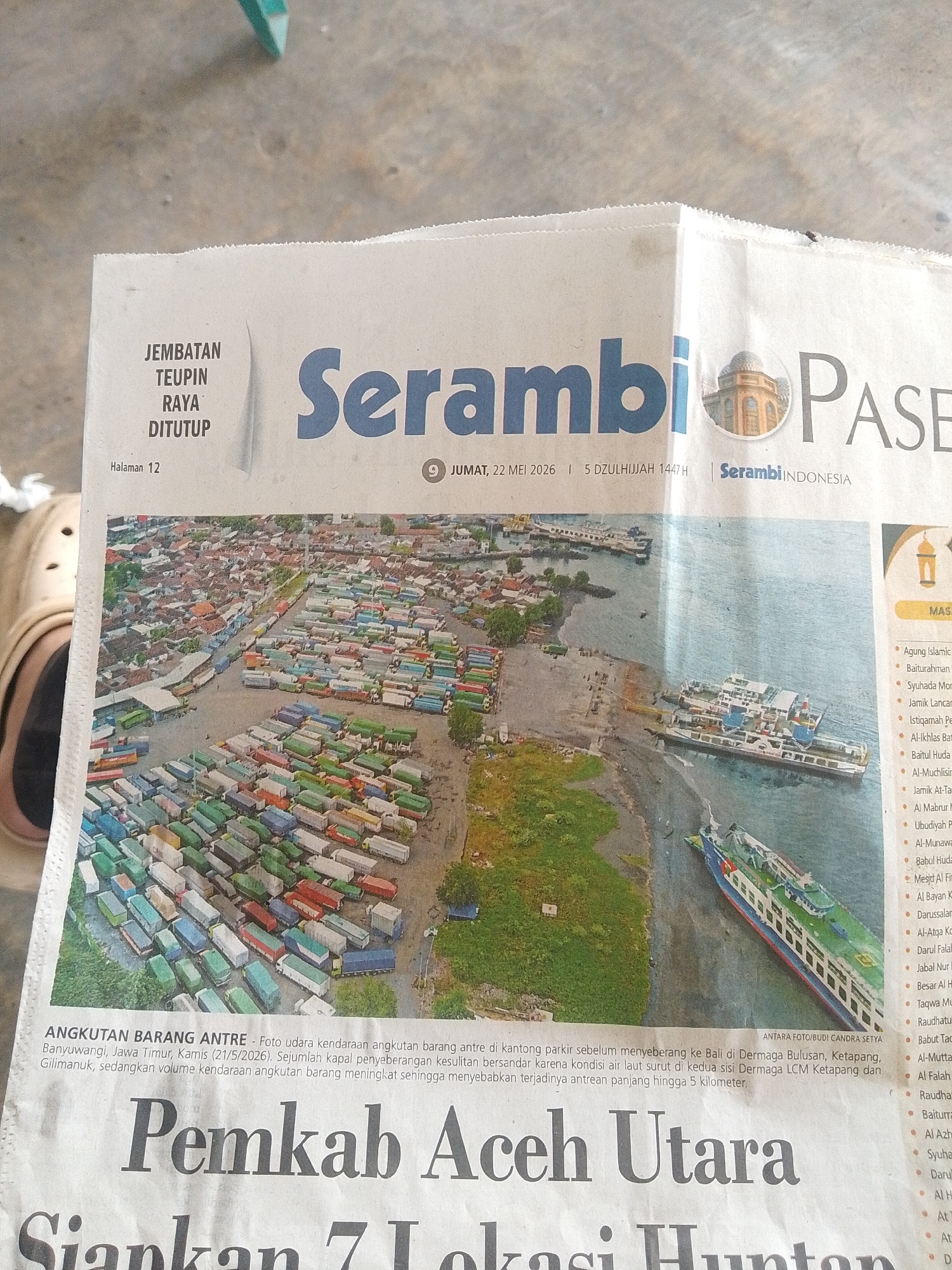 This is an image of a newspaper article with the headline "Serambi PASE". The date is JUMAT, 22 MEI 2026. The article, titled "ANGKUTAN BARANG ANTRE", shows an aerial photograph of a parking area congested with trucks waiting to cross over to Bali at Dermaga Bulusan, Ketapang, Banyuwangi, Jawa Timur. The text states that ships are having difficulty docking due to low tide conditions on both sides of Dermaga LCM Ketapang and Gilimanuk. The truck volume is high, creating a 5-kilometer queue. The text "Pemkab Aceh Utara Siapkan 7 Lokasi Hunton" is also visible.